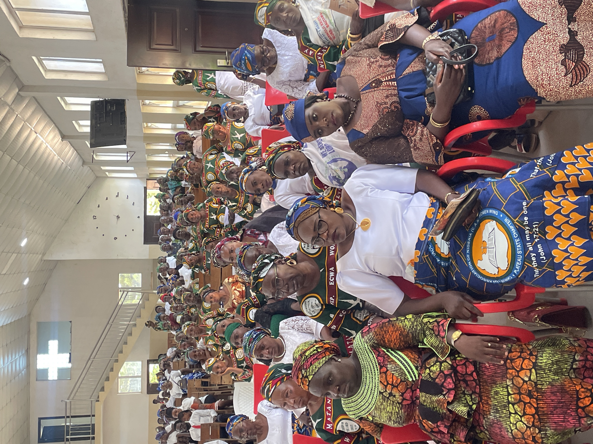 The image shows a large gathering of women inside a building, likely a church, in Bukuru, Nigeria. The women are seated in rows of red chairs and are dressed in colorful traditional attire, with many wearing headscarves. The building has a staircase to a higher floor and windows allowing natural light. A loudspeaker is visible on the left wall. Many of the women are wearing clothing with symbols and inscriptions indicating affiliation with a Christian association. Some of the inscriptions mention the Evangelical Church Winning All (ECWA) and the Christian Association of Nigeria (CAN) Plateau State Chapter. A cross-shaped window is also visible. The event appears to be a religious gathering.