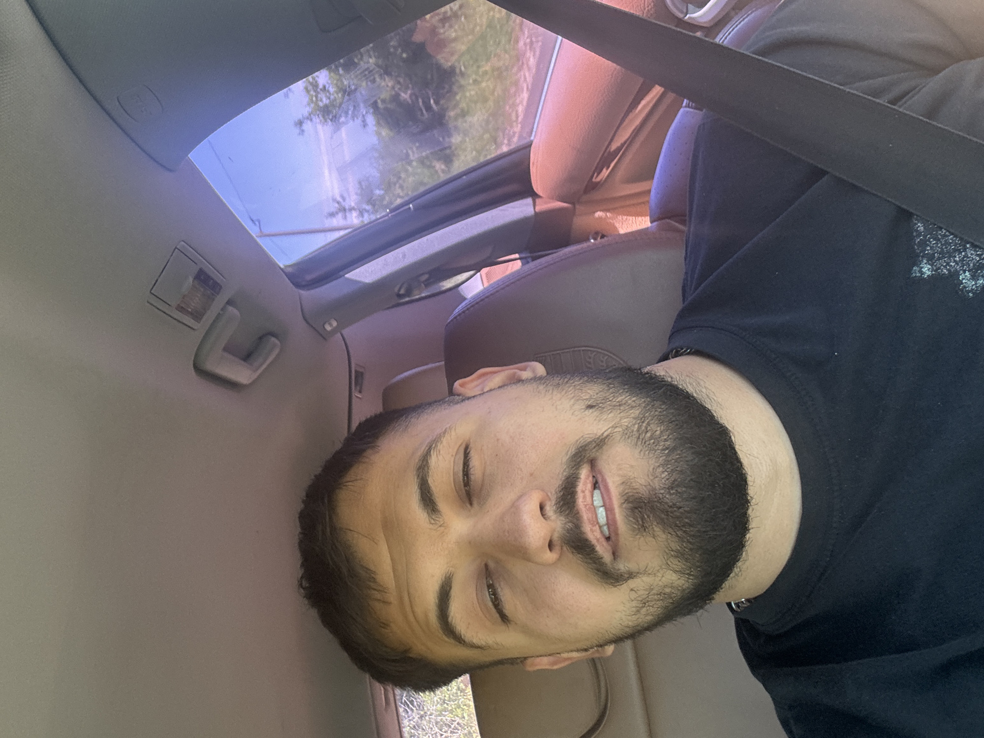 A man with a beard is sitting in a car, wearing a seatbelt and a black t-shirt. He is looking slightly downward and smiling with his eyes nearly closed. The car's interior is light grey, with brown leather seats. The sunroof is open, revealing a view of greenery outside. The location is Senovo, Bulgaria.
