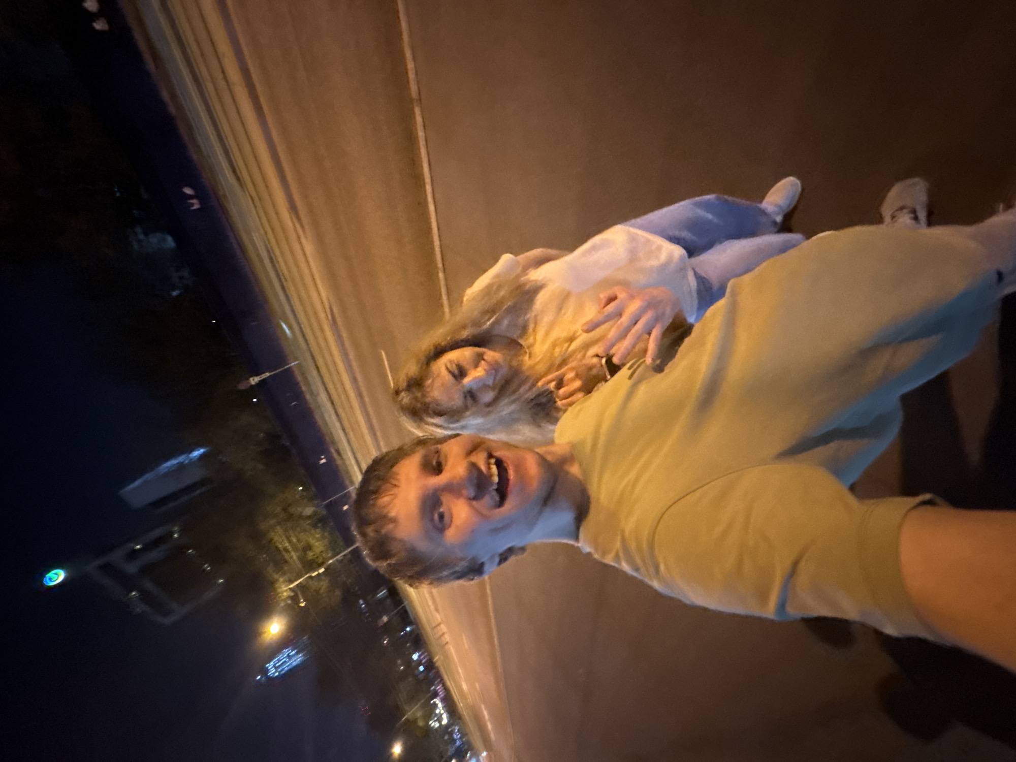 A selfie captures a man and a woman standing on a bridge at night in Moscow, Russia. The man, with light skin and hair, is smiling broadly at the camera. He is wearing a light green shirt. The woman, behind him and slightly to his left, has light skin, long blonde hair, and is wearing a white top and blue pants. They are embracing. In the background, the cityscape is visible with numerous lights. The bridge and the surrounding areas are well-lit.