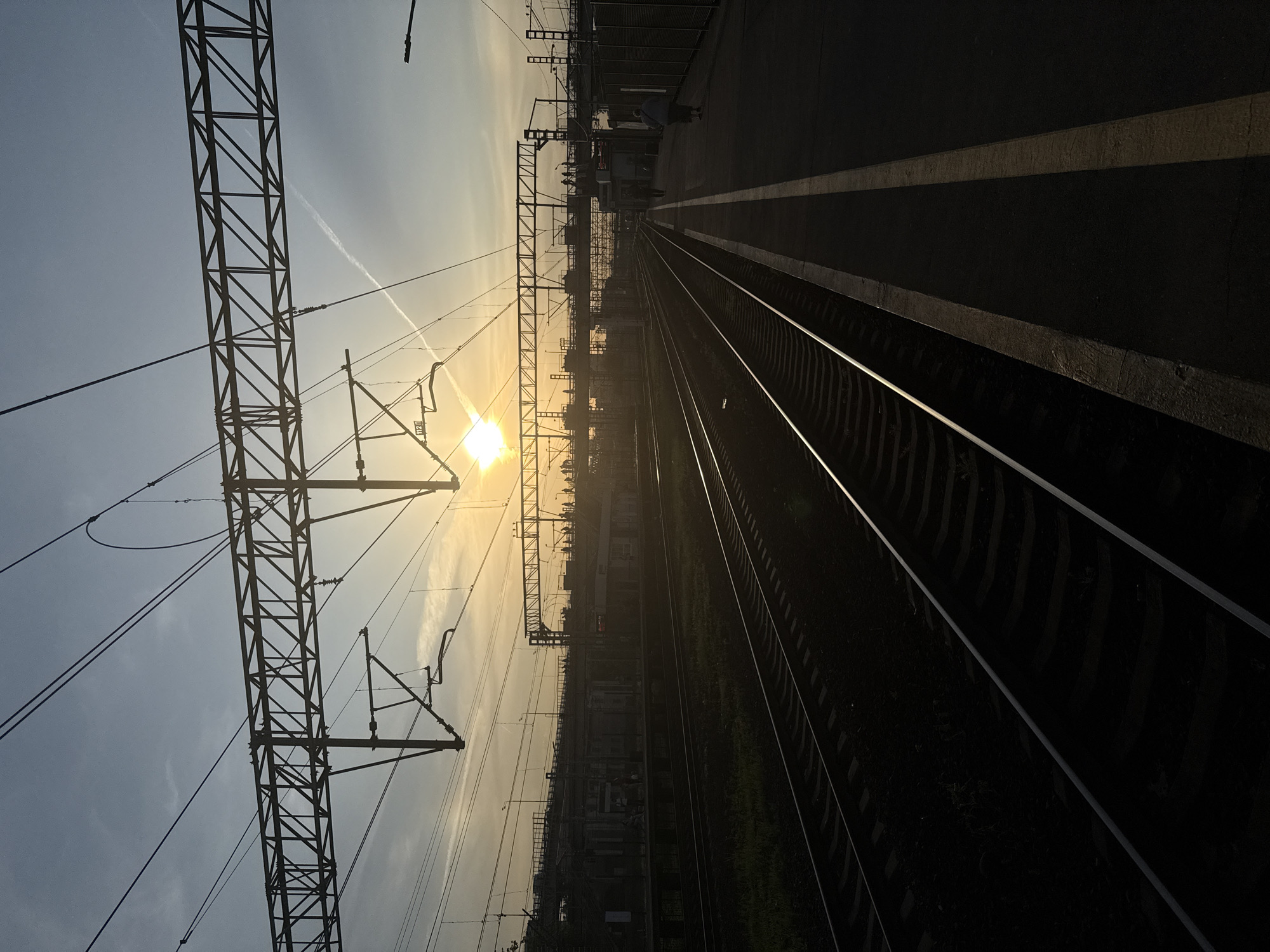 The image shows a railway track in Moscow, Russia, at sunset. The sun is visible between the tracks, creating a silhouette effect on the surrounding objects. The tracks run parallel to each other and converge in the distance. On the left side of the image, there is a metal structure supporting the overhead electrical wires for the trains. On the right side, there is a platform with two white lines indicating the edge. The platform is dark, and the tracks are illuminated by the setting sun. The sky is hazy, and a contrail is visible. In the distance, some buildings and vegetation can be discerned.