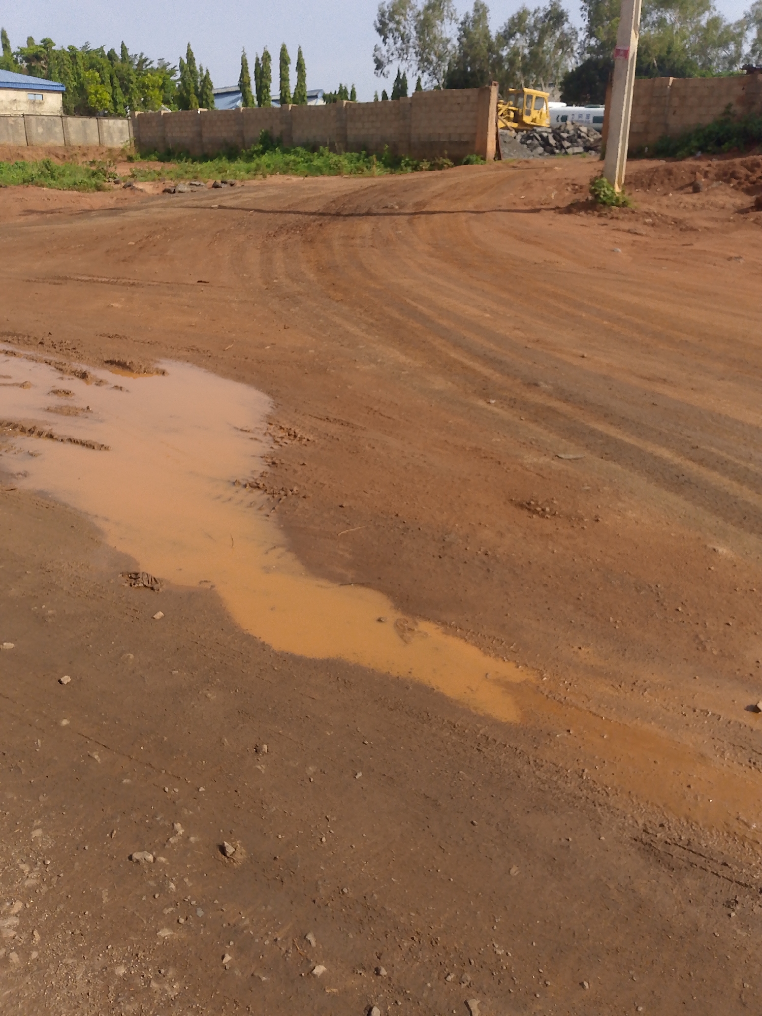 A construction site in Keffi, Nigeria, shows a muddy road with tire tracks and a large puddle of muddy water in the foreground. In the background, a brick wall separates the site from buildings and trees. A yellow bulldozer is visible behind the wall, suggesting ongoing construction work. The sky is clear and bright.