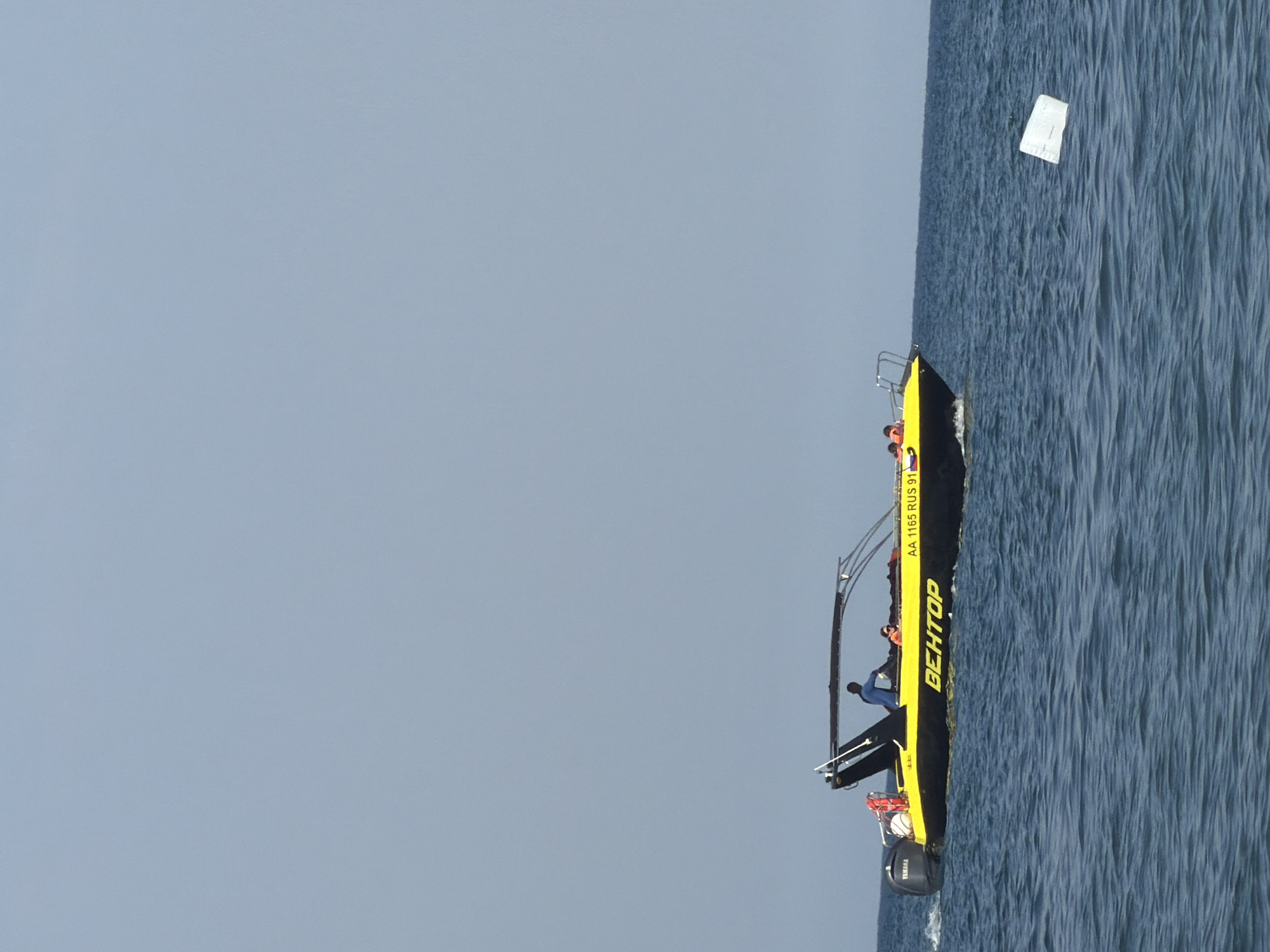 This is a photograph of a yellow and black boat on a body of water. The boat has a cabin and a railing. There are several people on the boat. One person appears to be wearing a blue shirt and black pants, and another is in an orange life jacket. There is also a Yamaha engine visible on the back of the boat. The boat has the word "BEHTOP" printed on its side in large, yellow letters. Above this word, there is smaller text that reads "AA 1165 RUS 91". In the water near the boat, there is a white, rectangular object, possibly a piece of debris or a marker. The water is choppy and appears to be deep blue. The sky is a pale, overcast blue. The orientation of the image suggests the photograph was taken with the camera held vertically. The scene appears to be taking place during the daytime, under cloudy weather conditions. The location context provided is Partenit, Ukraine.
