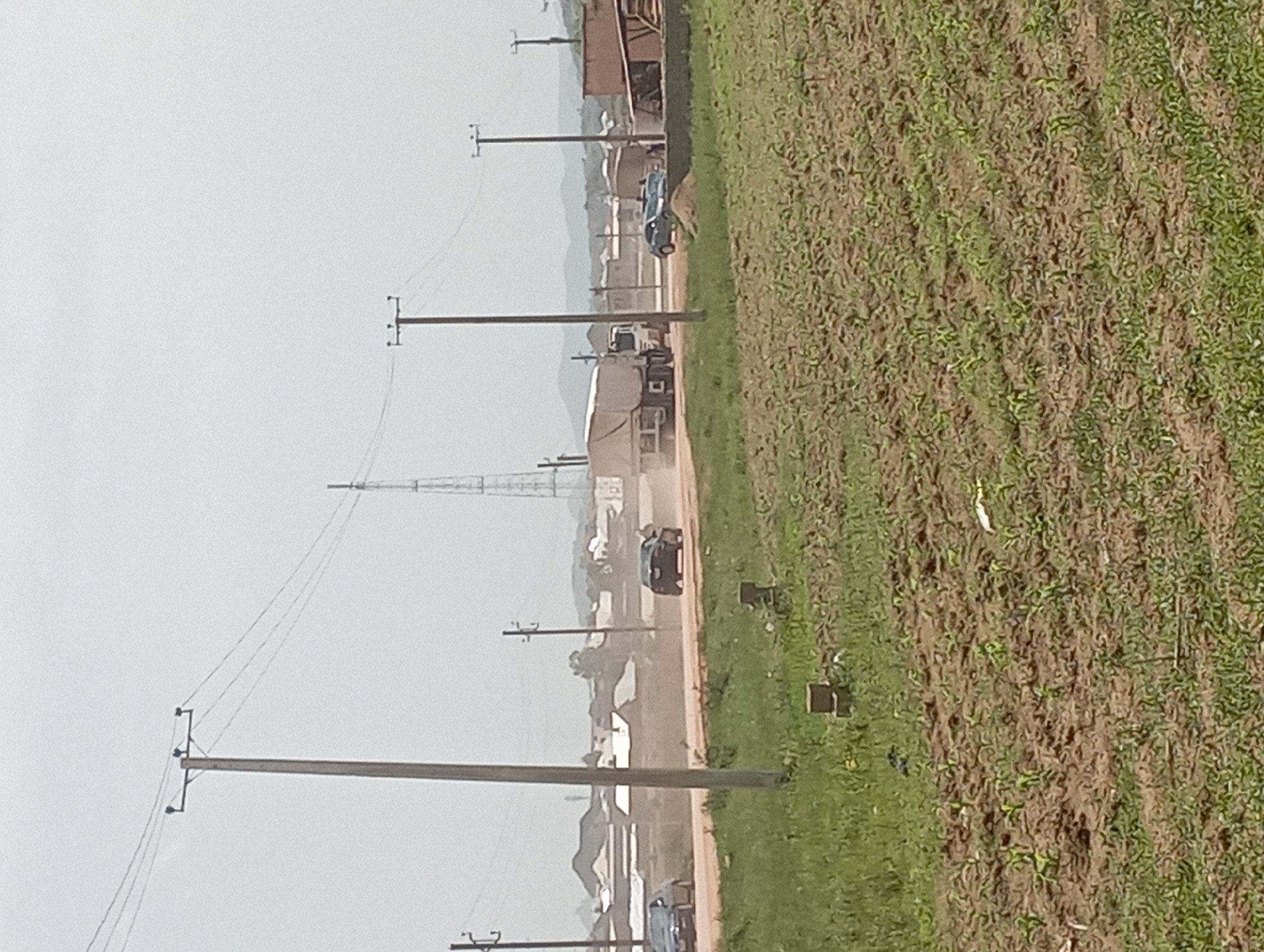 The image depicts a rural scene on a dusty road, likely in the outskirts of Jos, Nigeria. In the foreground, a field of young crops is visible, with rows of green sprouts emerging from the tilled earth. Beyond this field, a dirt road stretches across the landscape, with several vehicles traveling on it. To the left, a car is seen approaching, its headlights illuminated. Further down the road, a larger truck with a covered trailer kicks up a significant amount of dust as it moves. A dark-colored sedan is also visible further in the distance. Several utility poles with wires crisscross the sky, indicating the presence of infrastructure. In the background, a collection of buildings with corrugated metal roofs suggests a small settlement or village. A tall communication tower stands prominently against the hazy sky, and distant hills form the backdrop of the scene. The overall atmosphere is one of activity and everyday life in a semi-arid environment.