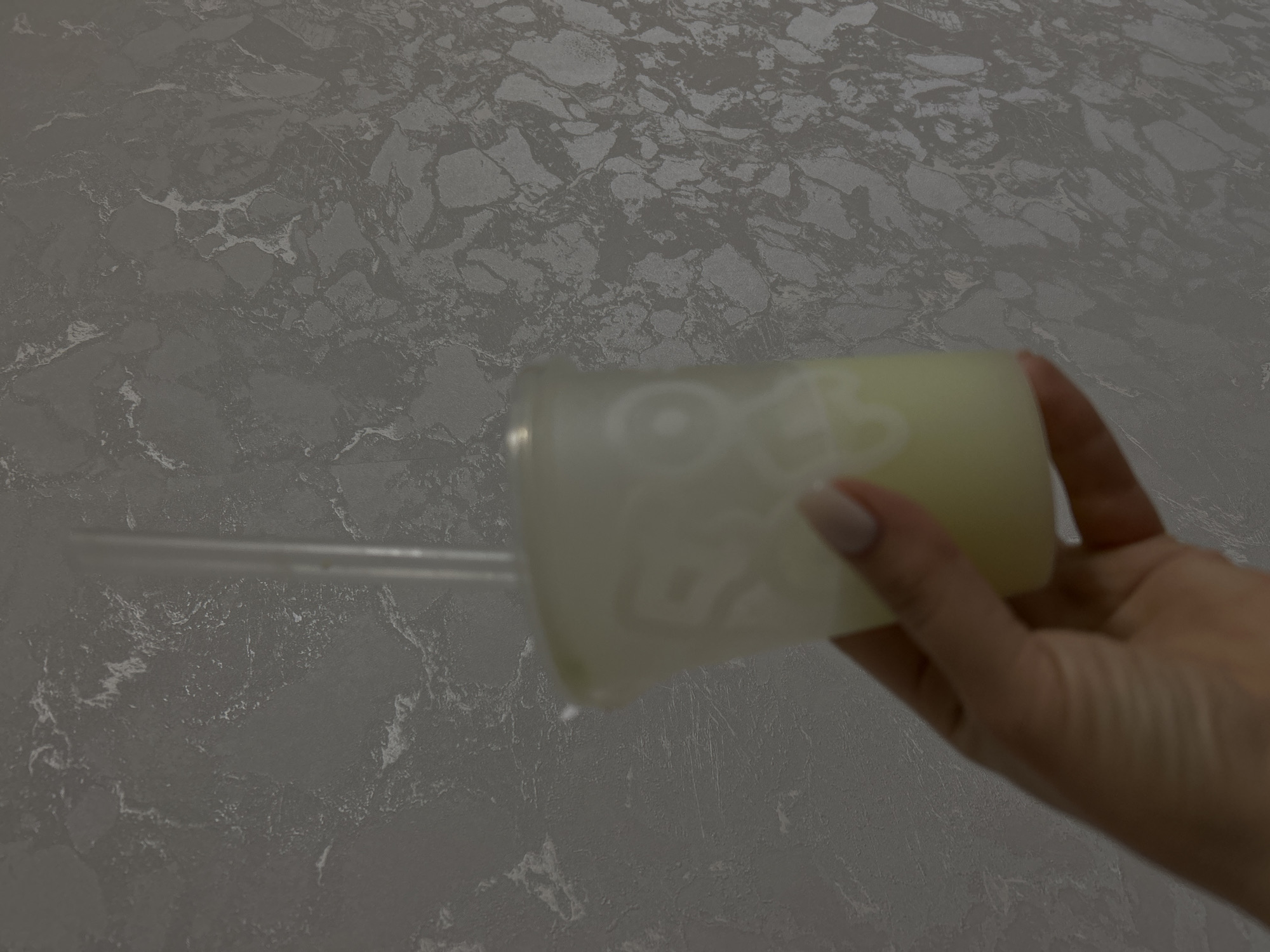 A person's hand is holding a clear plastic tumbler with a straw, filled with a yellow liquid. The tumbler has white lettering on it. The background is a grey surface with a stone-like pattern. Based on the text visible on the tumbler, it indicates the location is in the city of Chaikovsky, Russia.