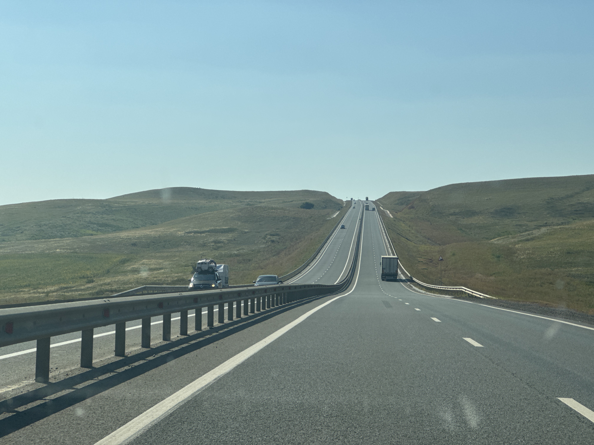 An asphalt highway stretches into the distance, curving and rising over rolling, grassy hills under a clear blue sky. A metal guardrail runs along the left side of the road. Several vehicles are visible on the highway: a white truck with cargo strapped to its roof, a dark sedan, and a white semi-trailer truck. The landscape is devoid of any structures or signs of habitation other than the road itself. The scene appears to be located in the open countryside. No people are discernible.