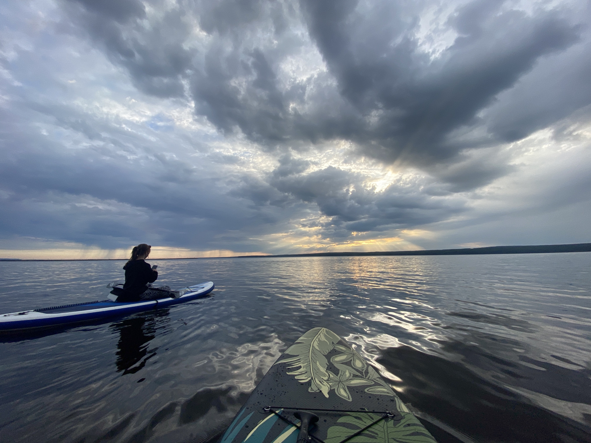 The image captures a tranquil outdoor scene of two stand-up paddleboarders on a wide body of water, likely a lake or reservoir, in Chaikovsky, Russia, during what appears to be late afternoon or early evening.
The perspective is from the bow of one paddleboard, looking towards a second paddleboard where a person is seated. This individual, seen from behind, wears a dark jacket and patterned leggings, and holds what appears to be a mobile phone. They are positioned on a blue and white inflatable stand-up paddleboard, with a paddle resting beside them. The paddleboard casts a clear reflection on the calm, dark water surface.
In the immediate foreground, the tip of the camera-side paddleboard is visible. This board features a distinctive dark green, black, and light green camouflage-like design with prominent leaf patterns. The water around both boards is rippled, catching and reflecting the light from the sky.
The sky is a dominant element, filled with dramatic, dark grey clouds that are broken in places by bright patches of sunlight. Striking crepuscular rays pierce through the clouds, illuminating the distant horizon and reflecting on the water. A faint, dark line of distant land or shoreline is visible beneath the bright horizon. The weather appears to be overcast but clearing, with the sun's rays indicating a beautiful, moody atmosphere. There is no visible text in the image.