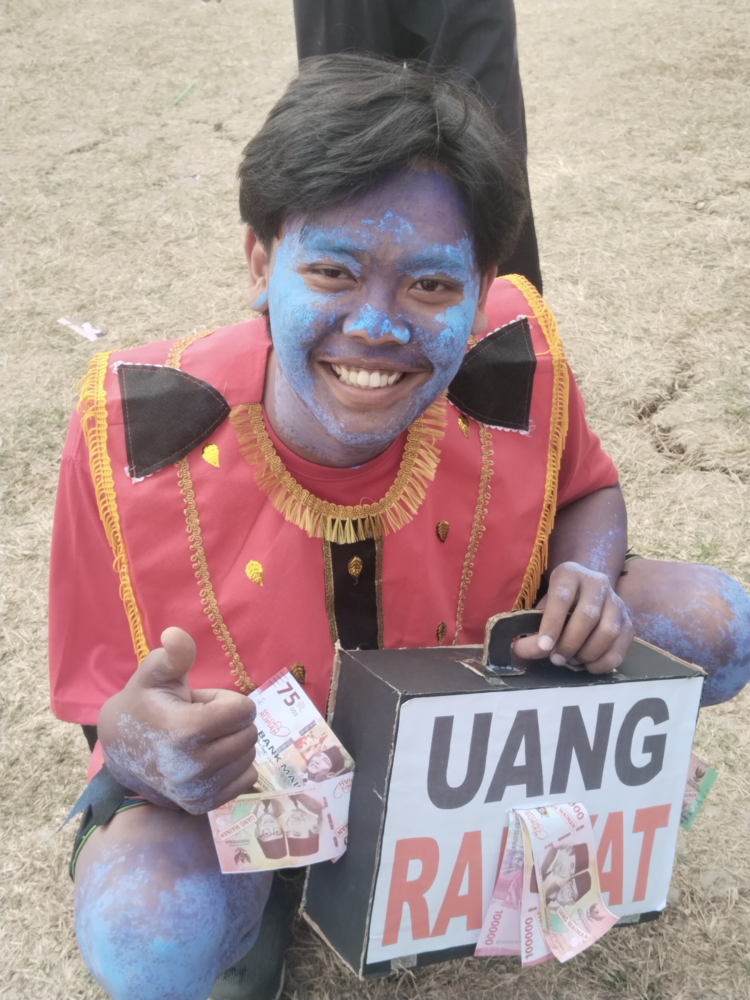 A young person with blue paint on their face and body, wearing a red and gold traditional outfit, is crouching on dry, grassy ground. They are holding a cardboard box with the words "UANG RAKYAT" (People's Money) written on it. Several Indonesian Rupiah banknotes are visible, some tucked into the box and others held by the person. The person is smiling and giving a thumbs-up gesture. The background consists of the dry, grassy terrain, possibly a field or park in the city of Pakulaut, Indonesia.
