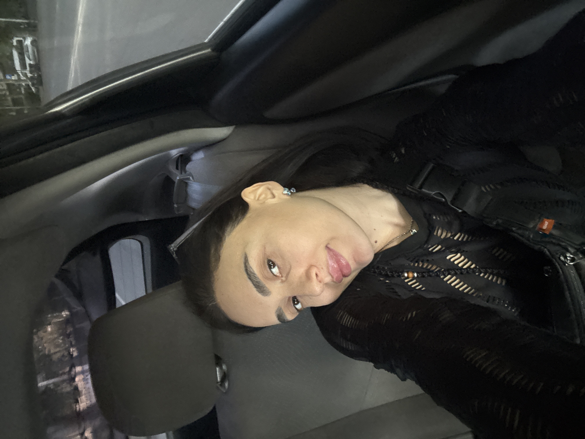 A woman is seated inside a car. She has dark hair and is wearing a black, semi-transparent top or jacket. The interior of the car is grey and black. Outside the car window, blurry lights suggest a city street at night. Based on the environment, the location is possibly in Tbilisi, Georgia.
