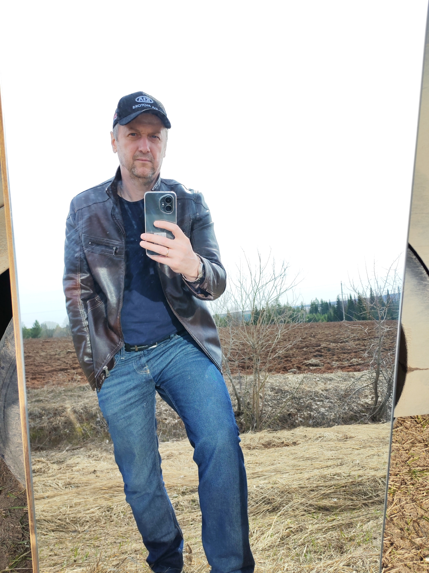 A middle-aged man is taking a selfie in a mirror, standing outdoors in a field. He wears a dark cap with the letters "ADA MOTORS" on it, a brown leather jacket over a navy blue shirt, blue jeans, and a belt. He is holding a smartphone with dual rear cameras. The background reflected in the mirror shows a rural landscape with brown fields, dry grass, bare trees, and a distant forest. The image was taken in Нечкино, Russia.