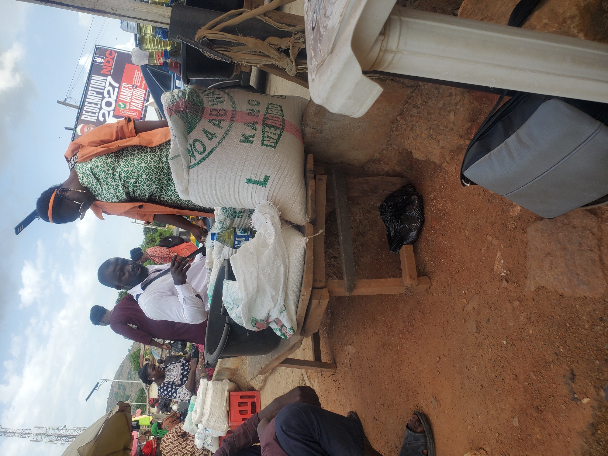 In Gwong, Nigeria, a busy market scene unfolds under a cloudy sky. A man in a white shirt is at the center, interacting with a woman in an orange jacket and a patterned green dress, who is looking down. They are near a wooden stall laden with sacks of produce and a black plastic basin. Several other people are visible in the background, some seated and engaged in market activities. The ground is dirt, with scattered stones. A large banner in the background advertises "Redemption 2027" with images and text in English and what appears to be a local language. Various items, including bottles of liquid, bags, and a red crate, are present in the scene, indicating a typical market environment.