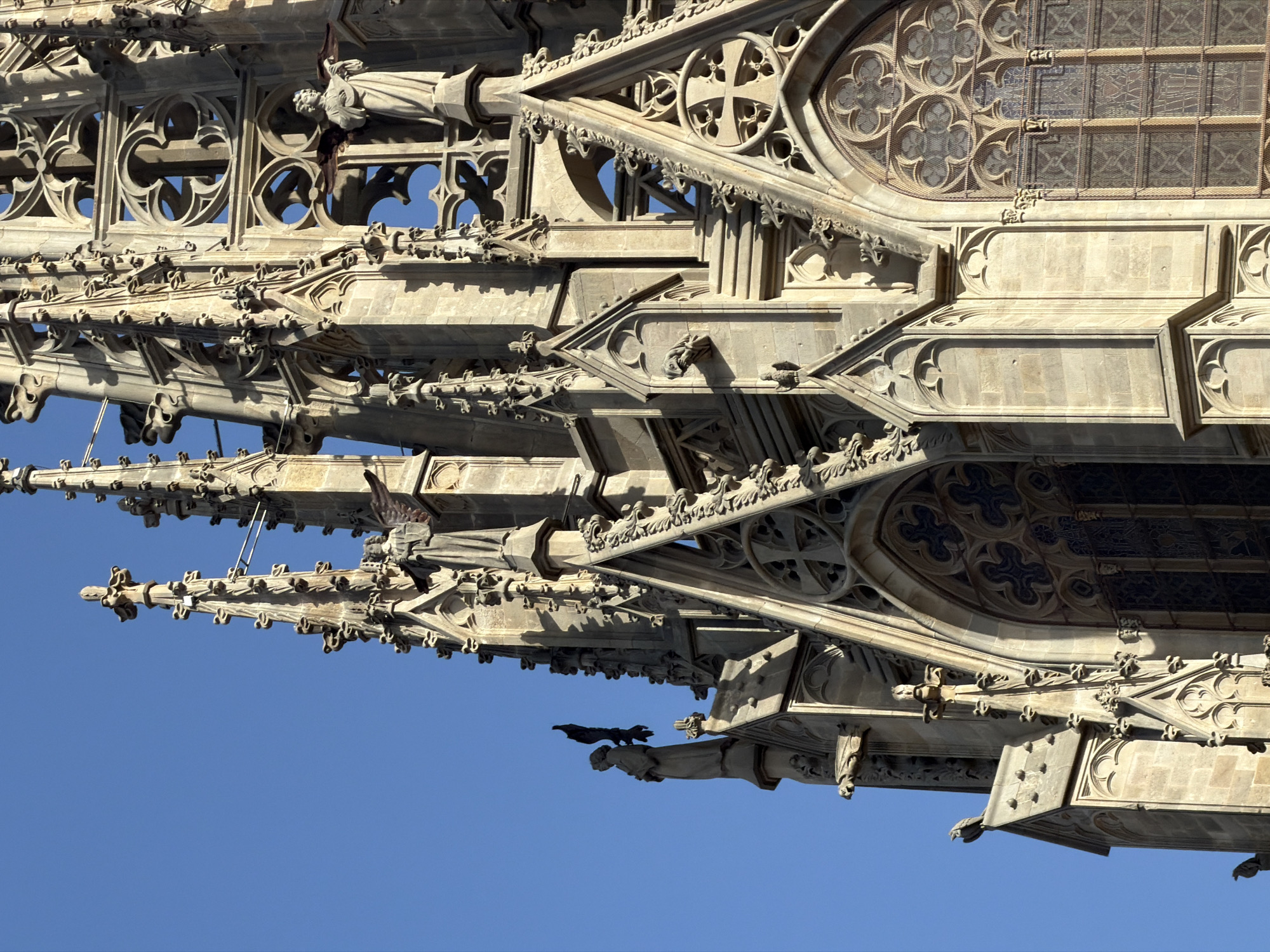 The image displays an upward view of the intricate stone architecture of a Gothic cathedral spire against a clear blue sky. The spire is adorned with numerous decorative elements including pointed arches, spires, gargoyles, and statues. Specifically, there is a statue of a person with wings, possibly an angel, on the right side of the image, and another figure of a person with outstretched arms on the left. Several birds are perched on the architectural features. The stonework appears aged and weathered. This is a detail of the Barcelona Cathedral, located in the Gothic Quarter of Barcelona, Spain.