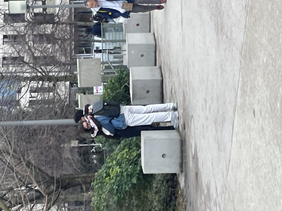 Two individuals stand close, one wearing a black tracksuit with white stripes on the sleeves, a blue denim jacket, and white pants. The other person wears a black jacket. They are near concrete cubes. Above them, an older person walks in the distance. Bare trees, bushes, and a building are visible in the background. The scene takes place on a gray paved surface, likely a sidewalk or plaza.