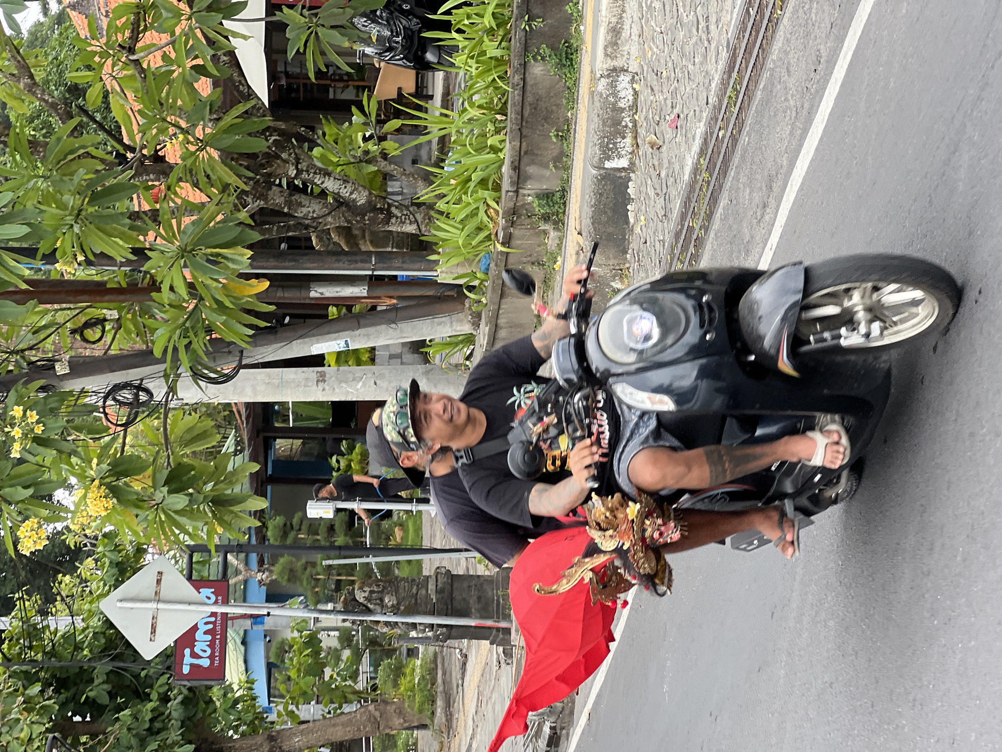 In Denpasar, Indonesia, two men ride a black scooter down a paved street lined with vegetation and buildings. The man in front is driving, wearing a black t-shirt, patterned baseball cap, and shorts. He has tattoos on his arms and a tattoo on his left leg. On the back of the scooter, a person is seated, wearing a black t-shirt and a cap. Attached to the back of the scooter is a large, folded red umbrella and a decorative object resembling a dragon head with gold accents. The scooter's headlight is illuminated. In the background, a sign for "Tamo Tea Room & Listening Bar" is visible. The street appears to be a public road with a sidewalk and some electrical wires overhead.