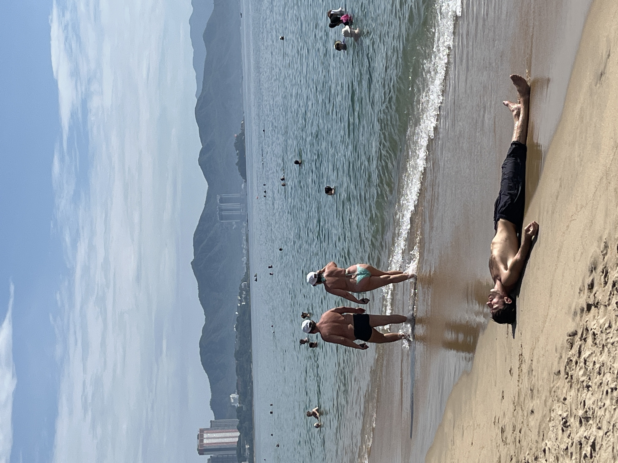 A man is lying supine on a sandy beach in Nha Trang, Vietnam, with his eyes closed and head turned to his right. He is wearing black swim trunks. Gentle waves are lapping at his feet. In the background, a couple walks into the water. Several other people are visible swimming and wading in the ocean. A line of mountains forms the horizon, with buildings and a coastline visible in the distance. The sky is bright blue with scattered clouds.