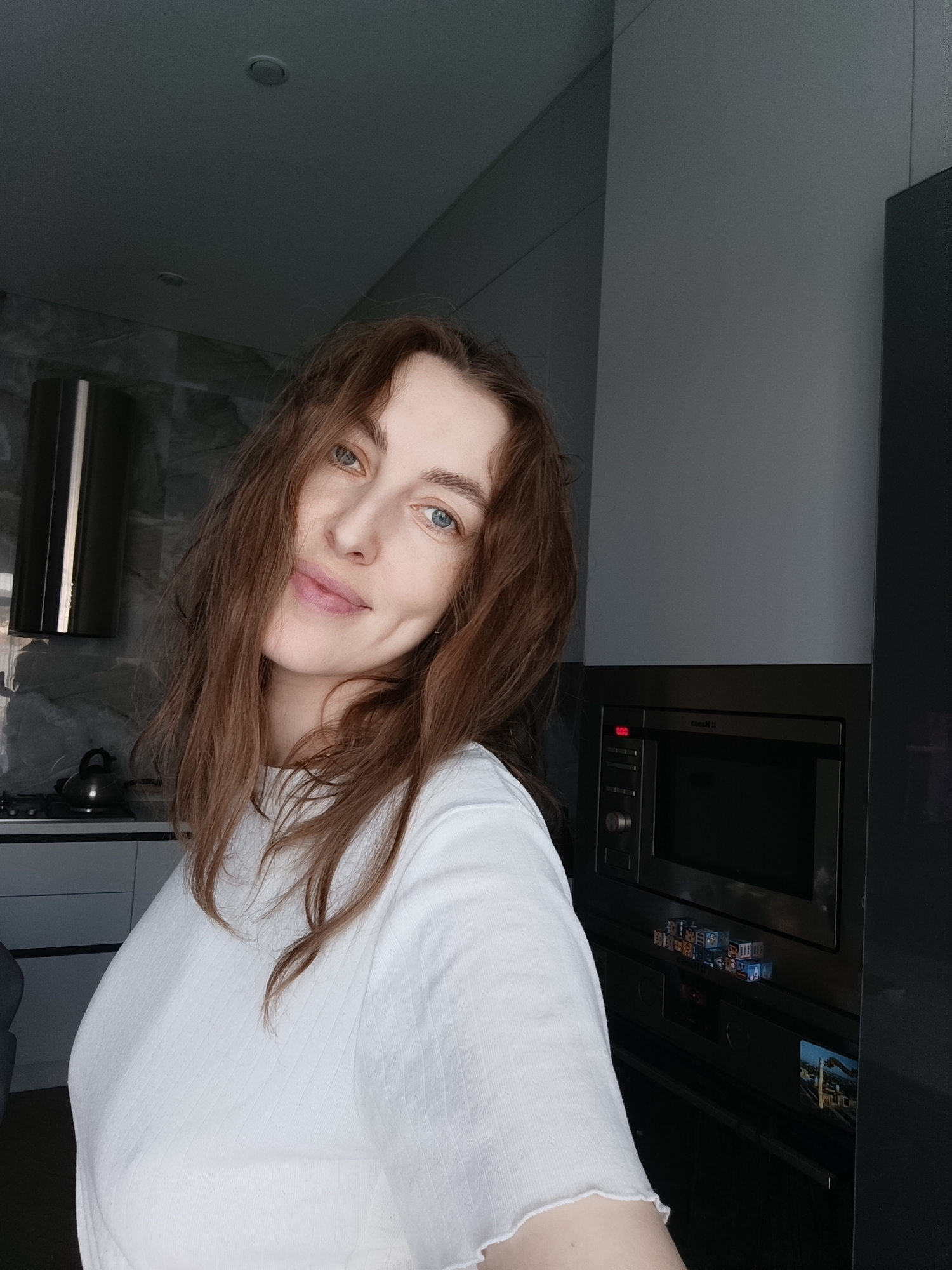 A young woman with wavy, reddish-brown hair and light blue eyes smiles gently, looking directly at the camera in a modern kitchen located in Chaikovsky, Russia. She wears a white, ribbed short-sleeved t-shirt, and a dimple is visible on her right cheek.
The background reveals a contemporary kitchen bathed in natural daylight. To the left, a sleek stainless steel range hood hangs above a gas stovetop, on which a black kettle rests. The wall behind the stovetop features a textured, grey, stone-like finish. The right side of the frame is dominated by dark grey cabinetry, integrating kitchen appliances. An eye-level microwave oven displays "0:00" in red and has a partially visible, indistinguishable brand name. Below it, small decorative cubes with various letters and symbols, including some Cyrillic characters (one appearing to spell "ПОЕЗД" - train), are stacked on the control panel of an integrated oven. A small rectangular image, possibly depicting a city or landscape scene, is affixed to the dark cabinet door to the right of the oven. The ceiling is light grey with recessed lighting. The woman appears relaxed and content.
