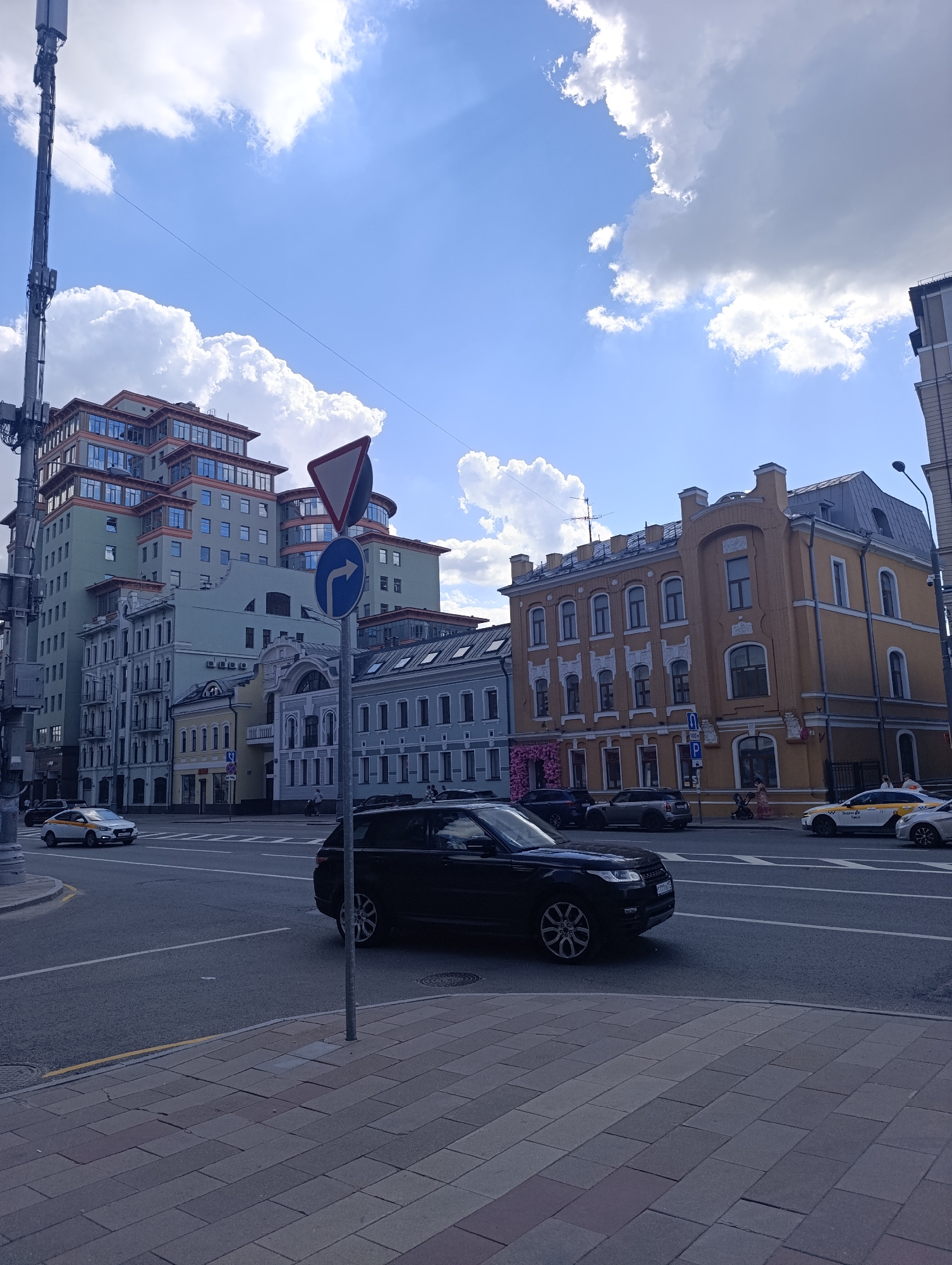 The image depicts a dynamic city street corner in Moscow, Russia, under a bright blue sky dotted with fluffy white clouds, indicating a clear and sunny day, likely during late morning or early afternoon. The scene is characterized by a blend of architectural styles and everyday urban activity.
On the left, a tall, multi-story modern building with a light green facade, numerous balconies, and a distinctive stepped or terraced design stands prominently. Beside it are several older, more ornate buildings with traditional detailing. To the right, a striking historical building with a yellow facade, elaborate white decorative trim around its windows and cornices, and a dark roof captures attention. A section of another blue-grey building is visible next to it.
The asphalt road is busy with vehicles. In the foreground, a black SUV, possibly a Range Rover, is seen in motion. Further down the street, two white and yellow taxis are visible, one being a Hyundai Solaris, both marked with "Tакси Go" (Taxi Go) and Russian lettering, common in Moscow. Other cars are present, parked or moving through the intersection. A tall cellular tower or mast extends vertically on the far left.
Street furniture includes various traffic signs: a triangular yield sign and a blue circular sign indicating a right turn ahead. A blue parking sign with the letter "P" and "100м" (100 meters) is also visible near the yellow building. The sidewalks are paved with grey and reddish-brown blocks. A few pedestrians are observed walking on the sidewalks, including one person pushing a stroller near the yellow building, contributing to the sense of a normal, active urban environment. The license plate on the black SUV appears to start with "Р111".