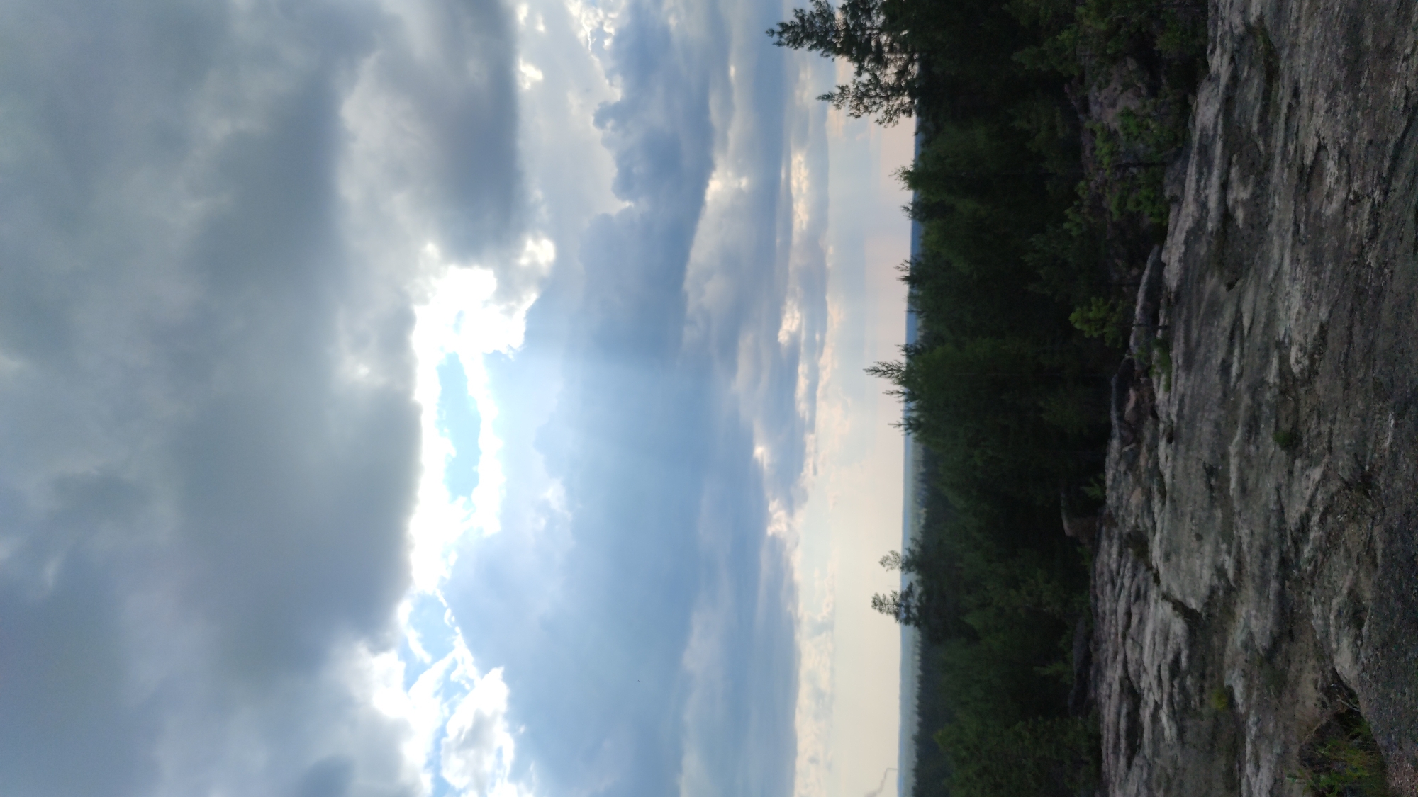 The image depicts a rocky, elevated landscape overlooking a dense coniferous forest under a dramatic, cloud-filled sky. The foreground is dominated by a rough, uneven surface of light gray and beige rock formations, with patches of sparse green vegetation. In the middle ground, a vast expanse of dark green pine trees stretches to the horizon, forming a thick forest canopy. Above the treeline, the sky is filled with heavy, dark gray clouds, punctuated by shafts of sunlight breaking through in places, creating a dramatic and atmospheric effect. The light suggests either early morning or late afternoon. The scene appears to be a natural, undeveloped area, possibly a viewpoint or a hiking trail, with no immediate signs of urban development or human presence within the frame. The location is not explicitly identifiable as Pitkyaranta, Russia, based solely on the visual elements present.