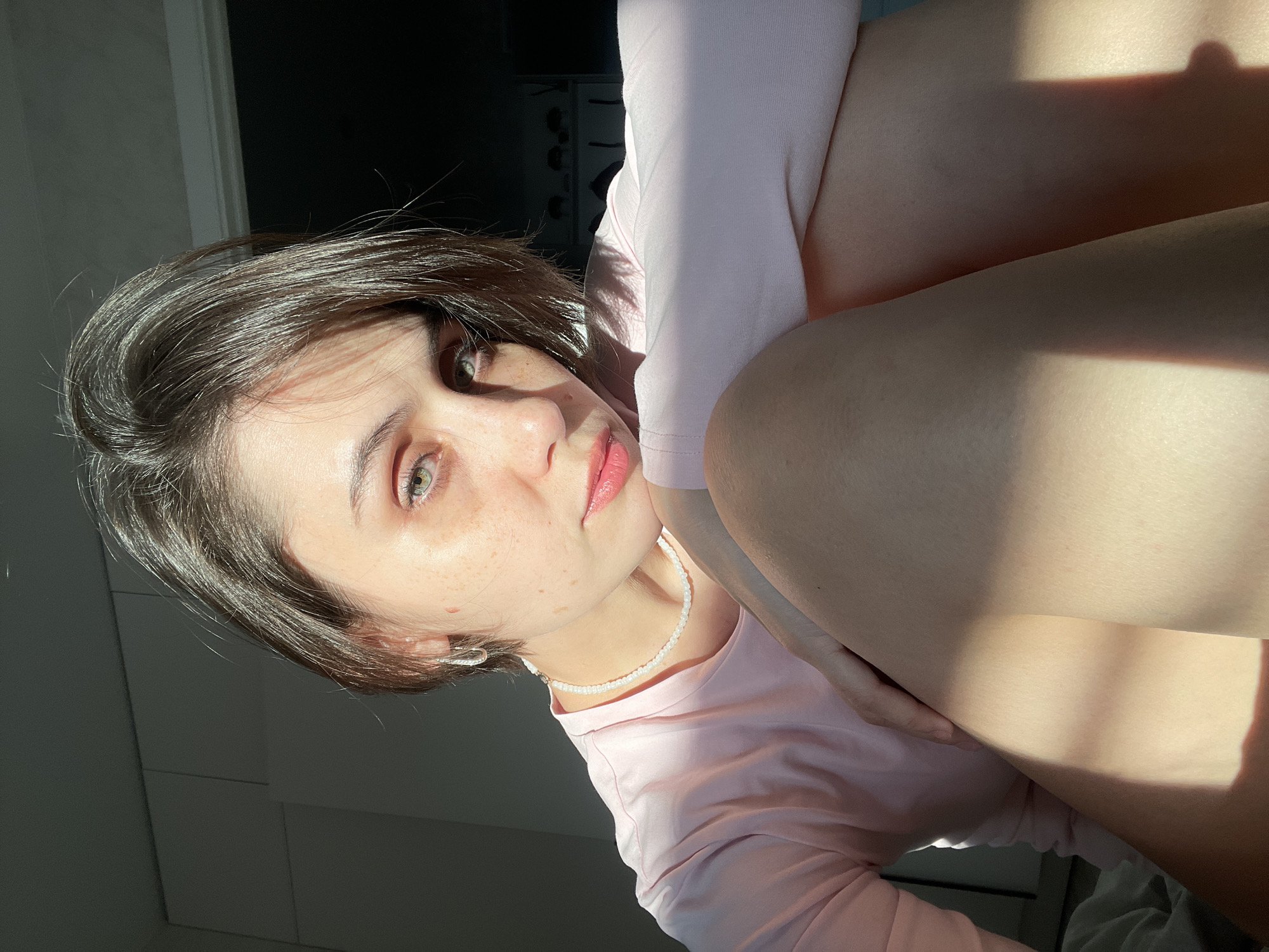 The image displays a female individual situated indoors, positioned in a crouched or seated posture with her knees bent and drawn upward, facing the viewer directly. She possesses short, dark brown hair, green eyes, a fair complexion with visible freckles on her nose and cheeks, and her lips appear to have a pink tint. She is dressed in a light pink long-sleeved shirt, a white beaded choker-style necklace, and a silver-colored hoop earring in her visible left ear. Her right arm is bent, with the forearm resting on or near her knees. Strong natural light, originating from the left, illuminates portions of her face, hair, chest, and bare legs, creating pronounced highlights and shadows across the scene. The immediate background to the left features light-colored wall panels. The background to the right is darker, showing a dark frame or molding at the top edge. No specific visual cues are present to determine the precise location within the city of Izhevsk, Russia.