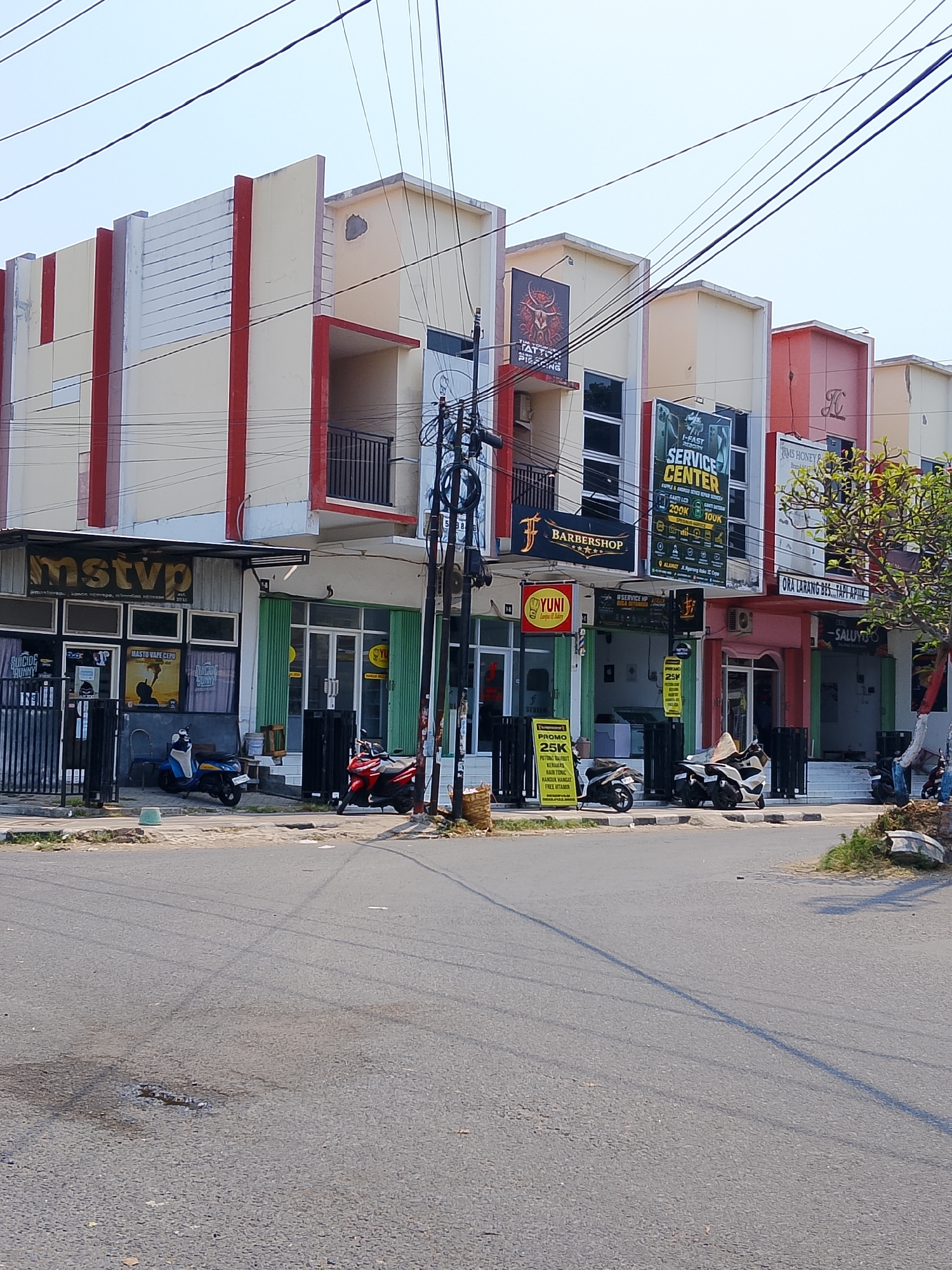 This is a daytime, eye-level, exterior, medium shot of a row of businesses on a street in Ngelo, Indonesia. The buildings are two-story structures painted in a cream and red color scheme with white accents. The foreground features an asphalt road with some water stains and tire marks. Several motorcycles are parked along the sidewalk in front of the businesses. In the background, power lines crisscross the sky, and a tree with green foliage is visible on the right side of the frame.
The businesses appear to be a mix of shops and service centers. From left to right, some of the visible signage includes "mstvp," "SUICIDE RUNNY," "i-FAST REBOOT SERVICE CENTER," "BARBERSHOP," and "YUNI." One sign prominently displays "SERVICE CENTER" with prices listed in Indonesian Rupiah (200K and 100K). Another sign advertises "PROMO 25K" with details about hair care and vitamin treatments. The overall impression is that of a commercial street in an urban or suburban area.