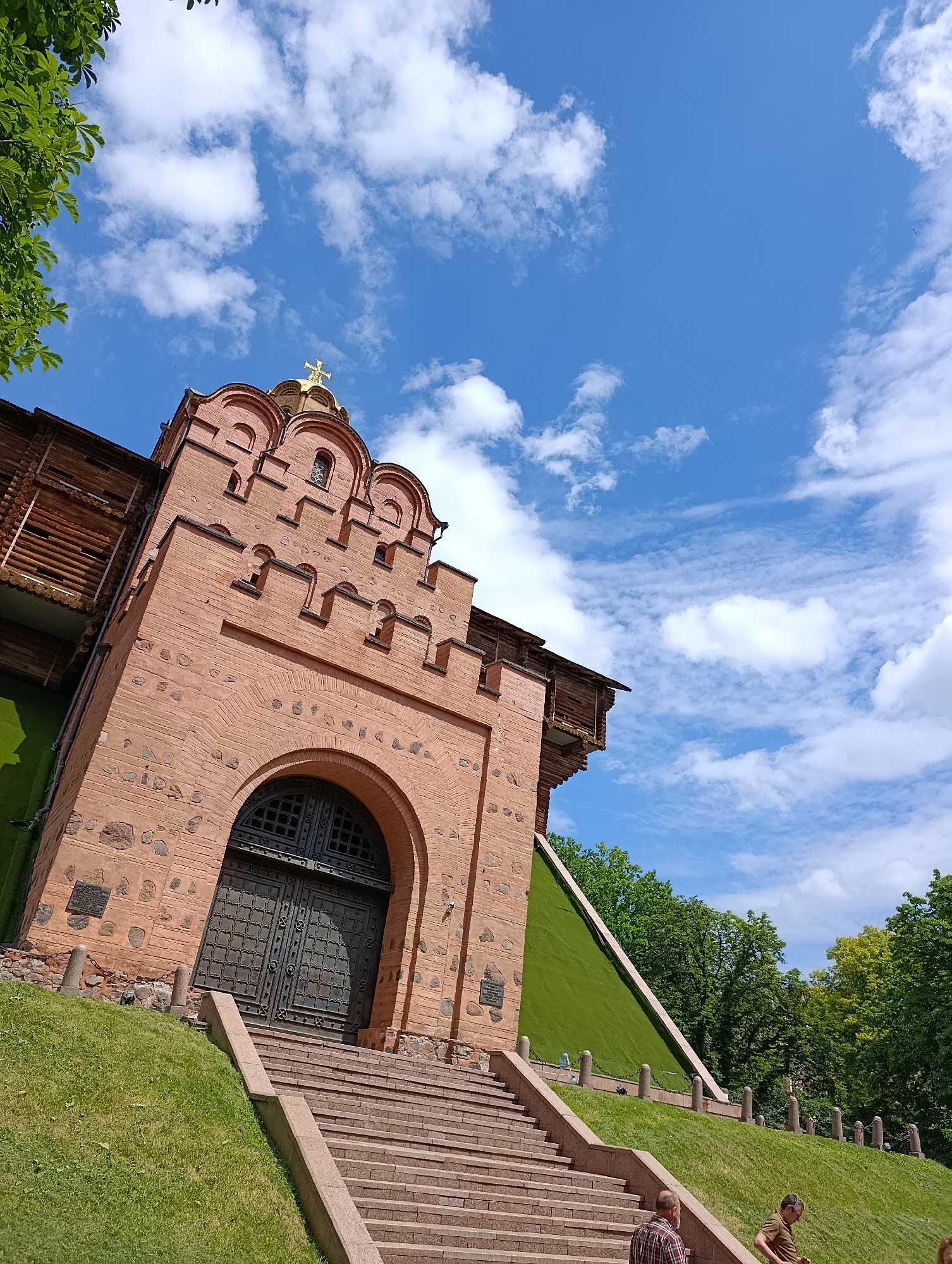 This image shows the Golden Gate of Kyiv, a historical monument in Ukraine. The gate is a massive brick structure with a large, ornate metal door. A wide stone staircase leads up to the entrance, and the gate is flanked by grassy slopes and trees. The sky is blue with fluffy white clouds, suggesting a pleasant day. A golden cross adorns the top of the gate, a religious symbol. The architecture appears ancient, hinting at the historical significance of the location. The image is taken from a low angle, emphasizing the grandeur of the structure. No visible text is present in the image.