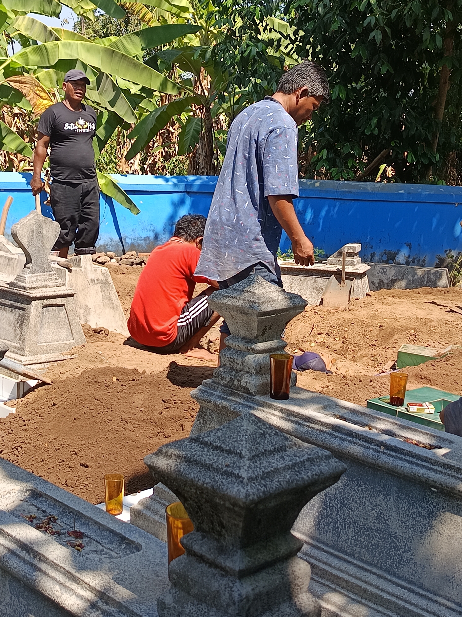 A group of men are engaged in the process of digging a grave at a cemetery in Geneng, Indonesia. The scene appears to be in the daytime, under bright sunlight, with lush green banana trees and other vegetation forming a natural backdrop. A vivid blue wall runs along the background, demarcating the cemetery grounds. In the foreground, gravestones and piles of excavated earth are visible. One man, wearing a black t-shirt and a cap, stands holding a shovel, seemingly overseeing the work. Another man, in a blue patterned shirt, is bent over, his head down, suggesting he is also involved in the digging. A third man, dressed in a red t-shirt and dark shorts with white stripes, is crouched low to the ground, actively working on the grave. Several amber-colored glasses, some filled with liquid, are placed on the gravestones, along with a pack of cigarettes. The overall impression is of a somber and laborious task being carried out in a rural, tropical setting.