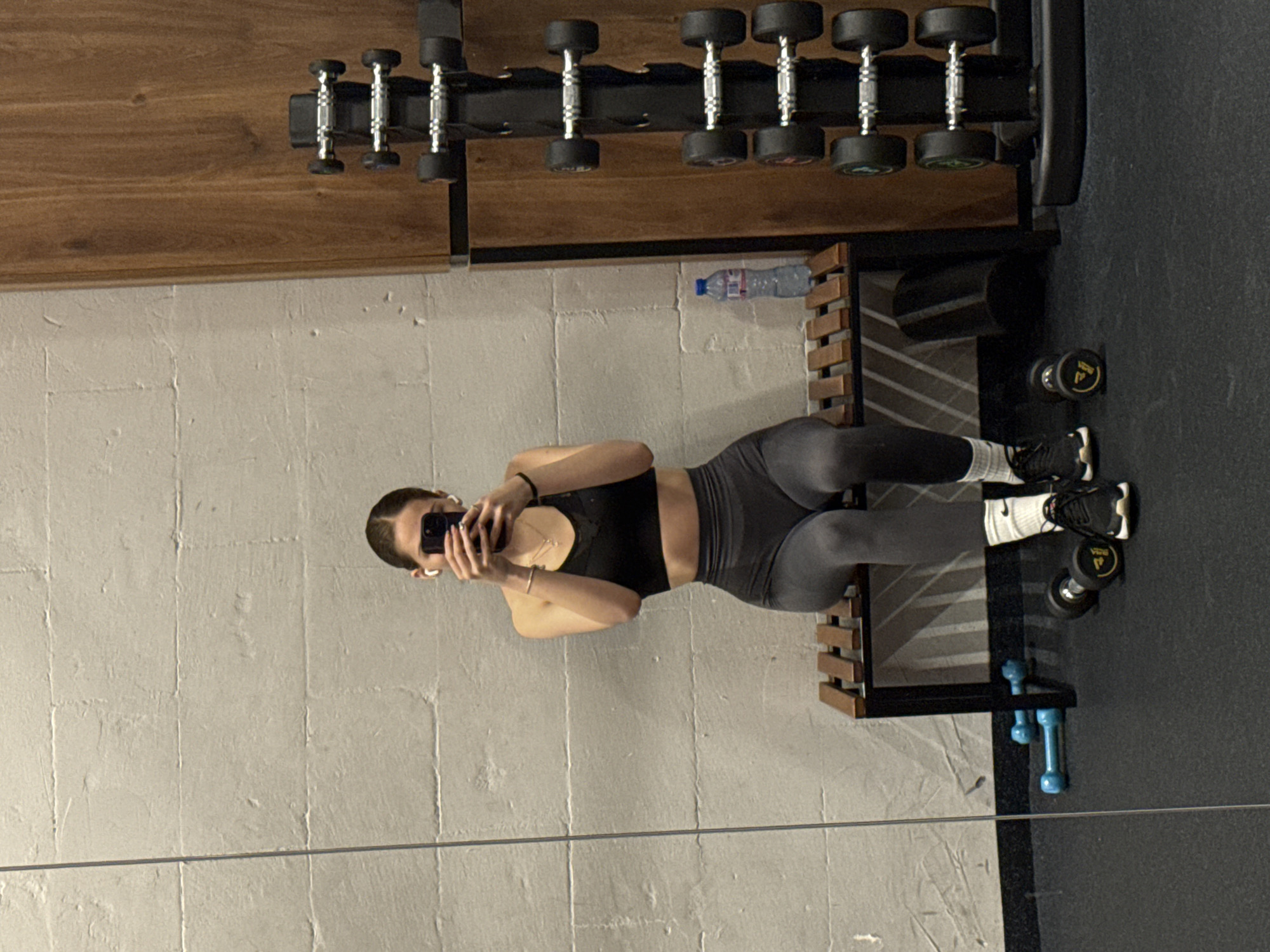 A woman is seen in a gym, reflected in a mirror. She is wearing athletic wear (black sports bra and grey leggings), white socks, and dark shoes. She is holding a smartphone in her left hand, apparently taking a selfie. The gym equipment visible in the background includes a dumbbell rack with various weights. There is also a water bottle placed on a wooden and black metal structure next to the dumbbell rack. The wall has a light-colored, textured surface. The location cannot be determined based on the image.