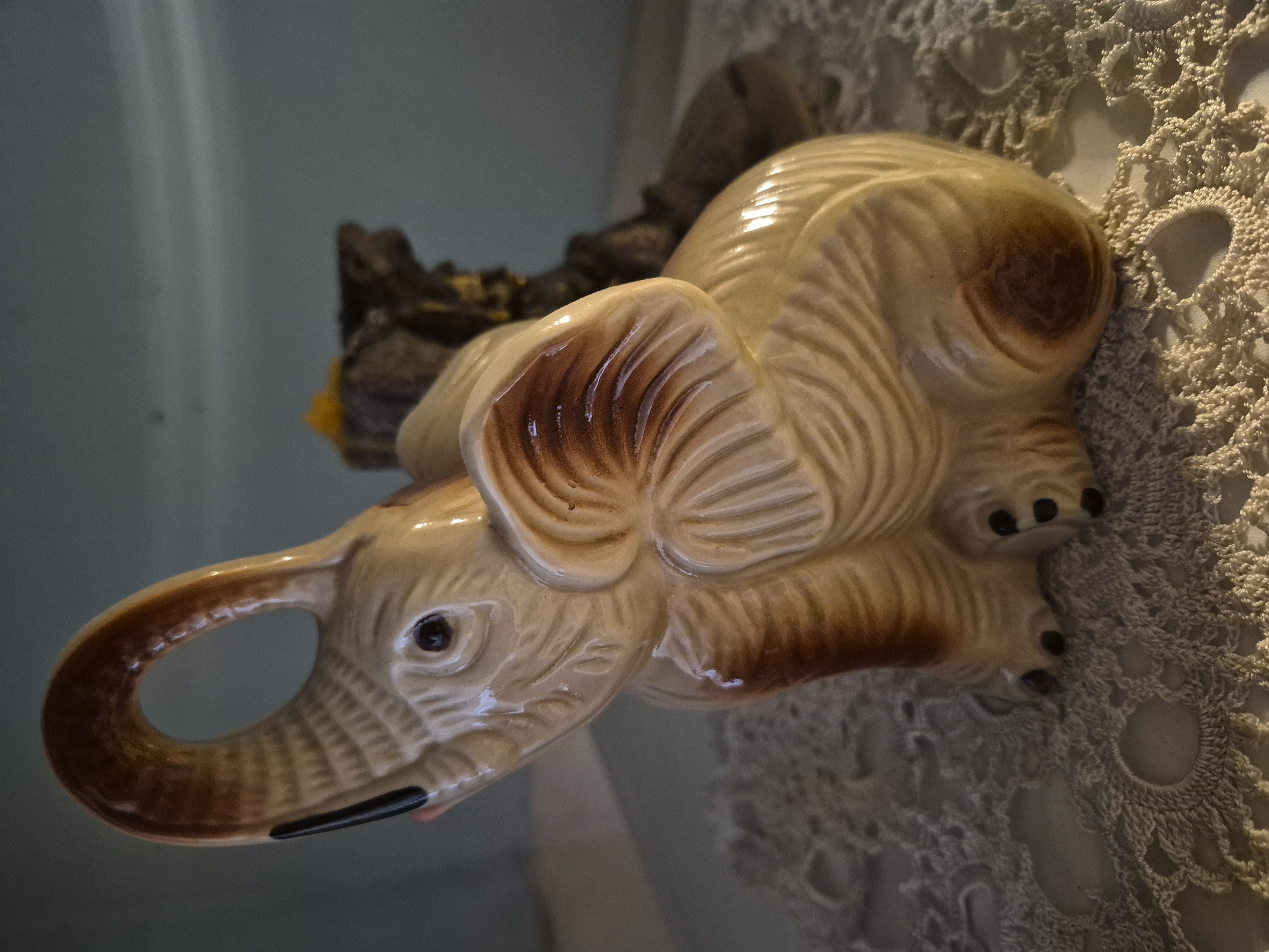 The image shows a ceramic figurine of an elephant with its trunk raised. The elephant is a light beige color with brown accents on its ears, trunk, and back. It is sitting on a white, crocheted doily. In the background, there is a blurred object that appears to be a rock formation with some yellow and brown elements. The lighting suggests an indoor setting, possibly a home or a decorative display. The scene is devoid of any specific geographical markers that would allow for pinpointing a location within Vanadzor, Armenia.