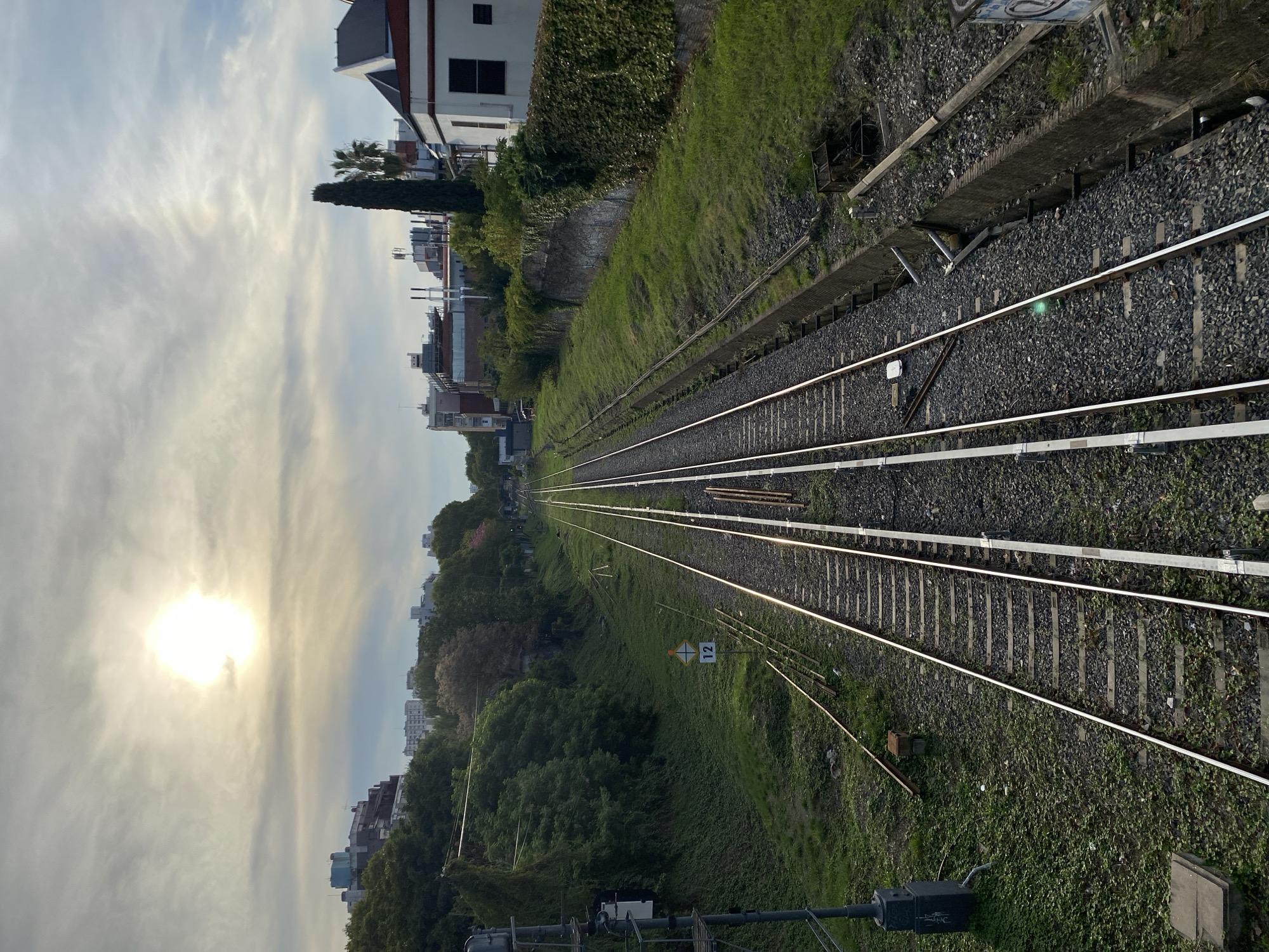 The image depicts a view along a railway track, receding into the distance towards a cityscape under a bright, cloudy sky. The tracks consist of two sets of rails laid on gravel, bordered by green vegetation on one side. In the background, buildings and trees make up the urban environment. A small sign with the number "12" is visible along the trackside. The sun is high in the sky, creating bright, diffuse light. The image does not provide sufficient evidence to definitively place the scene in Buenos Aires, Argentina.