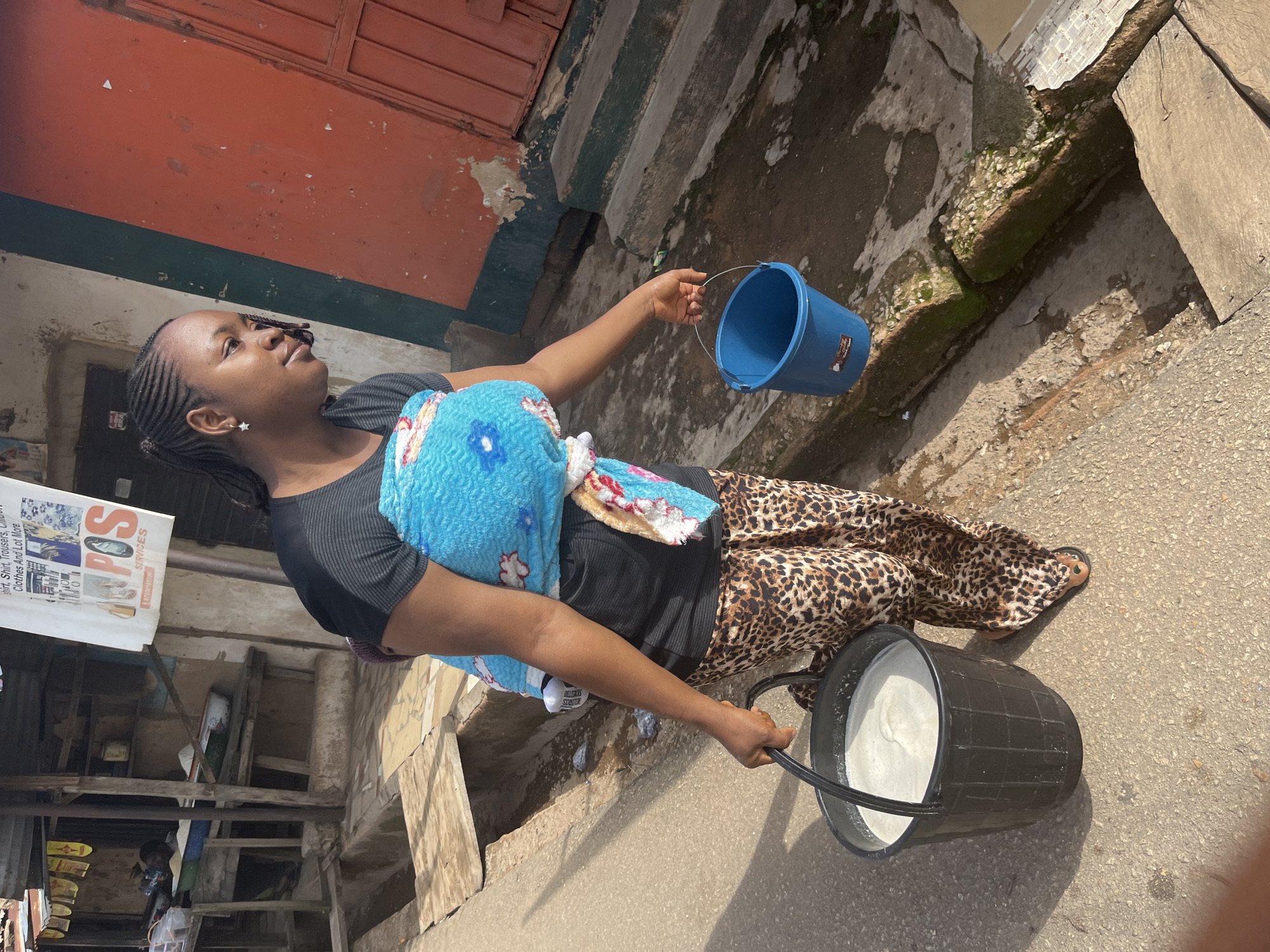 In Jos, Nigeria, a young woman carries two buckets, one filled with white liquid, while her child is wrapped in a blue blanket on her back. She wears a black t-shirt and patterned pants, and her hair is styled in braids. The scene unfolds on a street with a drainage ditch running alongside, suggesting a typical urban environment in Jos. The background shows a marketplace with stalls and buildings.