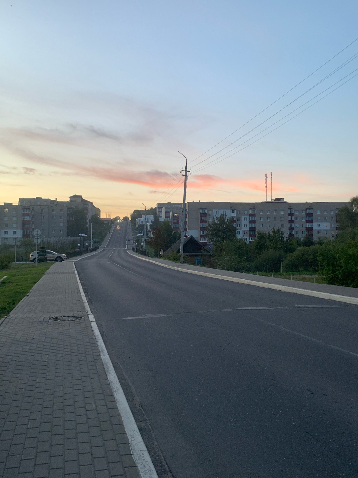 The image depicts a street scene in Kapyĺ, Belarus, during what appears to be sunset. A paved road with a white line marking the lane dominates the foreground and stretches into the distance, ascending a gentle incline. To the left, a grey brick sidewalk runs parallel to the road, featuring a manhole cover. Across the street, a line of residential buildings, typical of Soviet-era architecture, are visible, interspersed with trees and vegetation. One building on the right displays what appear to be cell towers on its roof. The sky exhibits soft hues of pink and orange from the setting sun, contrasting with the lighter blue above. A single car is parked on the left side of the road. The overall impression is one of a quiet, urban evening.