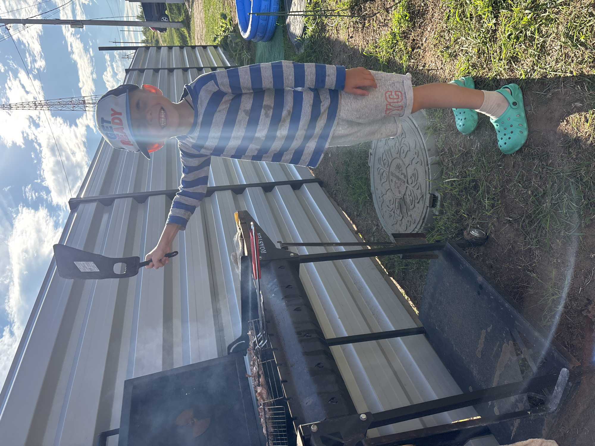 An outdoor shot shows a young boy standing next to a barbeque grill in what appears to be a backyard in Chaikovsky, Russia. The boy, wearing a blue and gray striped long-sleeve shirt, gray shorts with red lettering, and bright green clogs with tan socks, grins at the camera. He holds a black spatula in his right hand, poised above the grill. The grill, a dark metal construction with a rack of meat cooking inside, is positioned in front of a corrugated metal fence. Behind the boy, a blue inflatable pool and some greenery are visible. The sun is shining brightly, casting shadows on the ground. The overall scene suggests a casual, warm-weather activity.