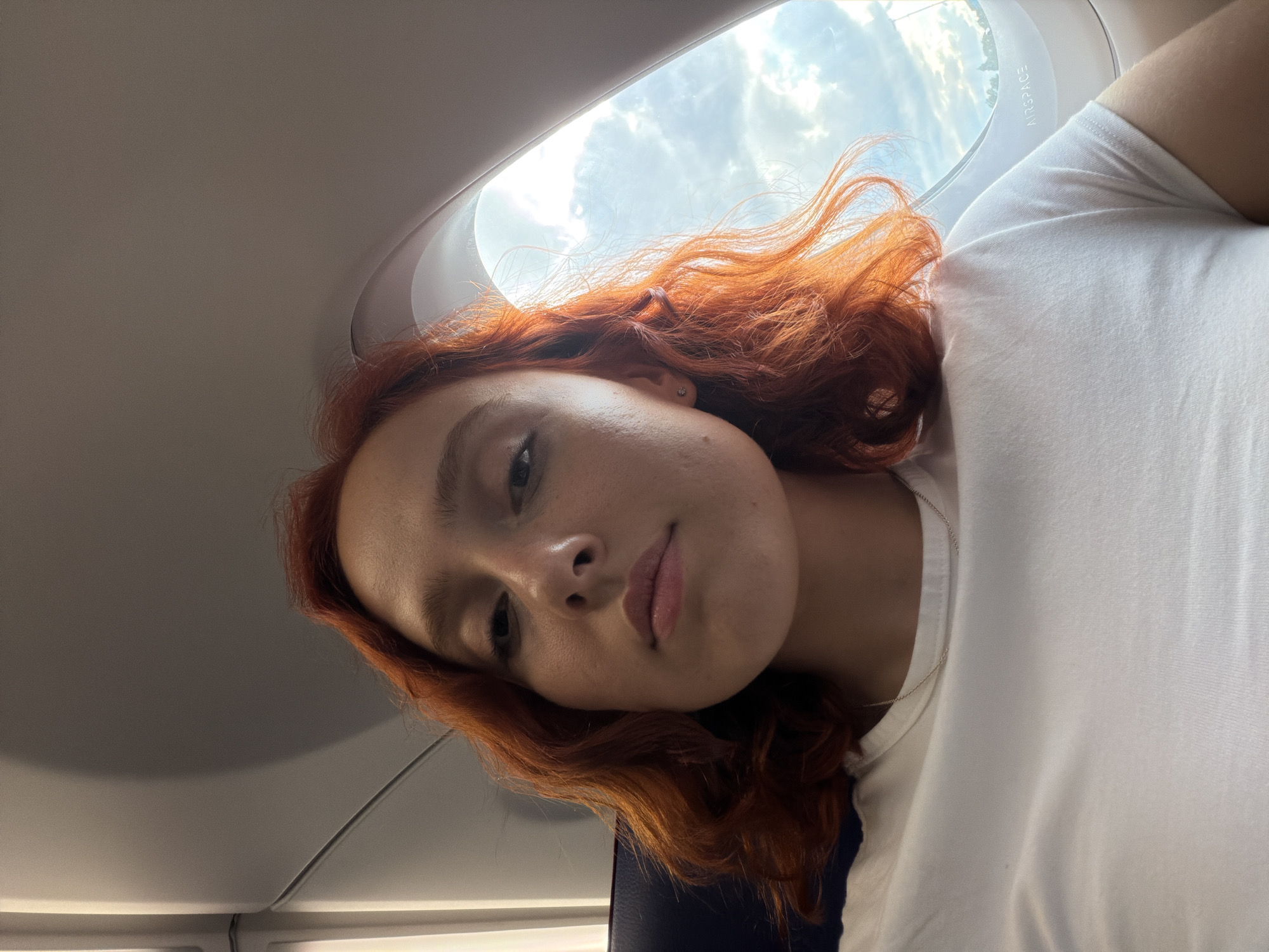 A young woman with vibrant red hair and fair skin is captured in a selfie taken from a window seat on an airplane. She is wearing a simple white t-shirt and a delicate silver necklace. Her hair is styled in loose waves that frame her face. The natural light from the airplane window illuminates her features, highlighting her serious expression. Outside the window, a cloudy sky suggests the plane is in flight. The interior of the airplane is visible, with the grey wall and curved window frame defining the space. The text "AIRSPACE" is subtly visible on the window frame. There is no indication of her location in Memmingen, Germany within the image.