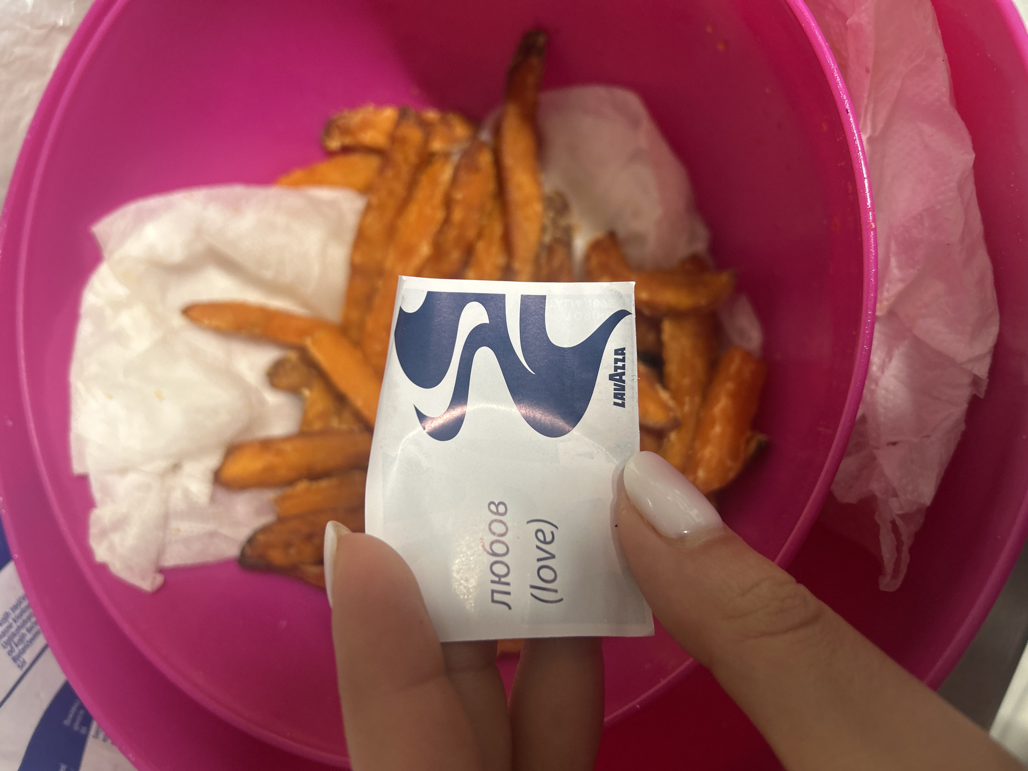 The image shows a pink bowl containing what appears to be sweet potato fries and a white paper towel. A hand is holding a Lavazza sugar packet with the word "любов" (love) written on it. The scene is indoors, likely in a home kitchen. Based on the sugar packet, the location may be in Bulgaria, specifically in Burgas.