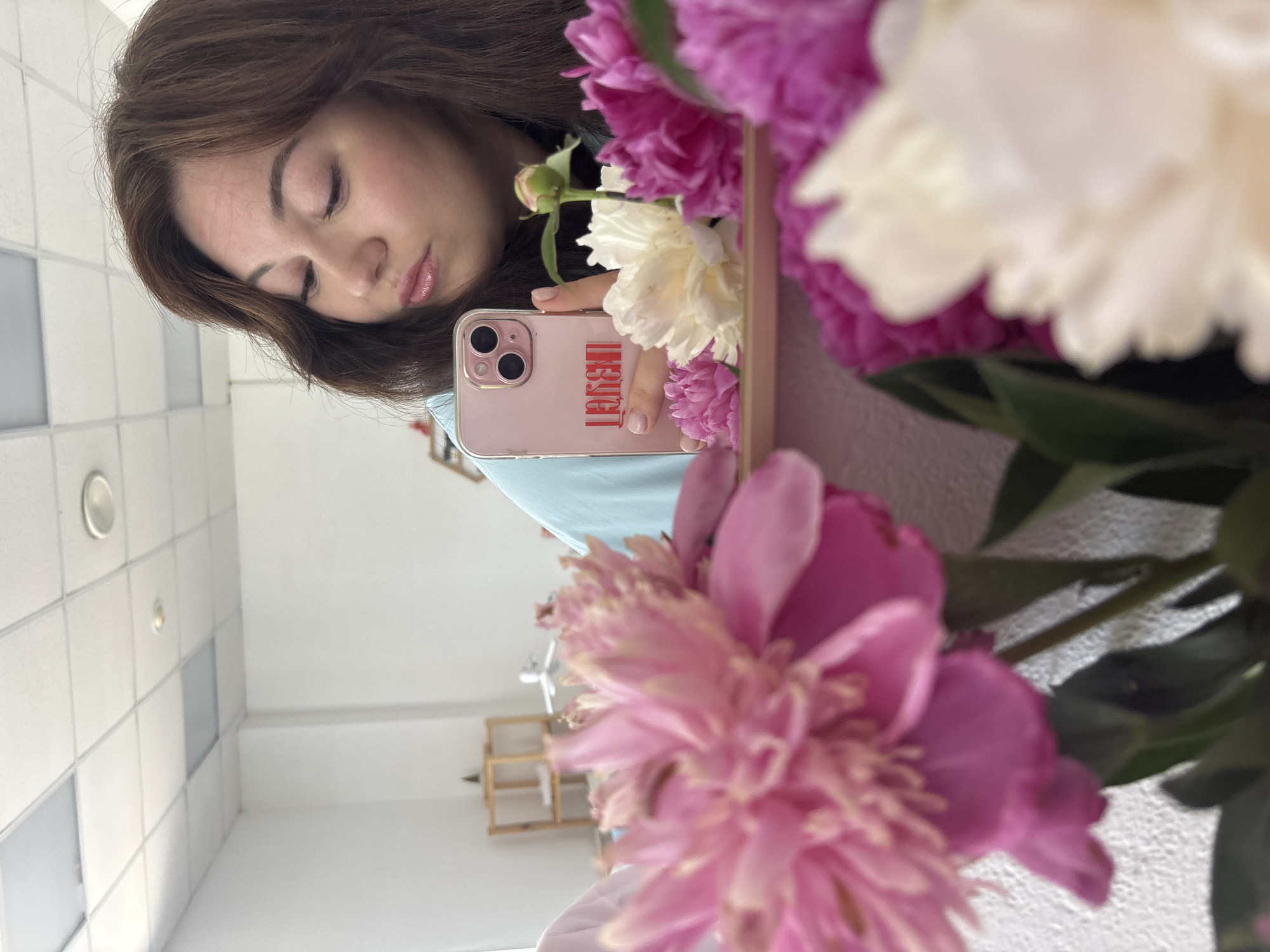 In an indoor setting, a young woman with dark brown hair is reflected in a mirror. She is holding a pink smartphone in her right hand, and the phone displays the red text "LOVE" in an artistic font, which is reversed due to the mirror. She is wearing a light blue garment. In the foreground, out of focus, are several large peonies in shades of pink and white. The background visible in the mirror shows a white wall with fluorescent ceiling lights and a shelf unit. There is no indication of the city of Chaikovsky, Russia.
