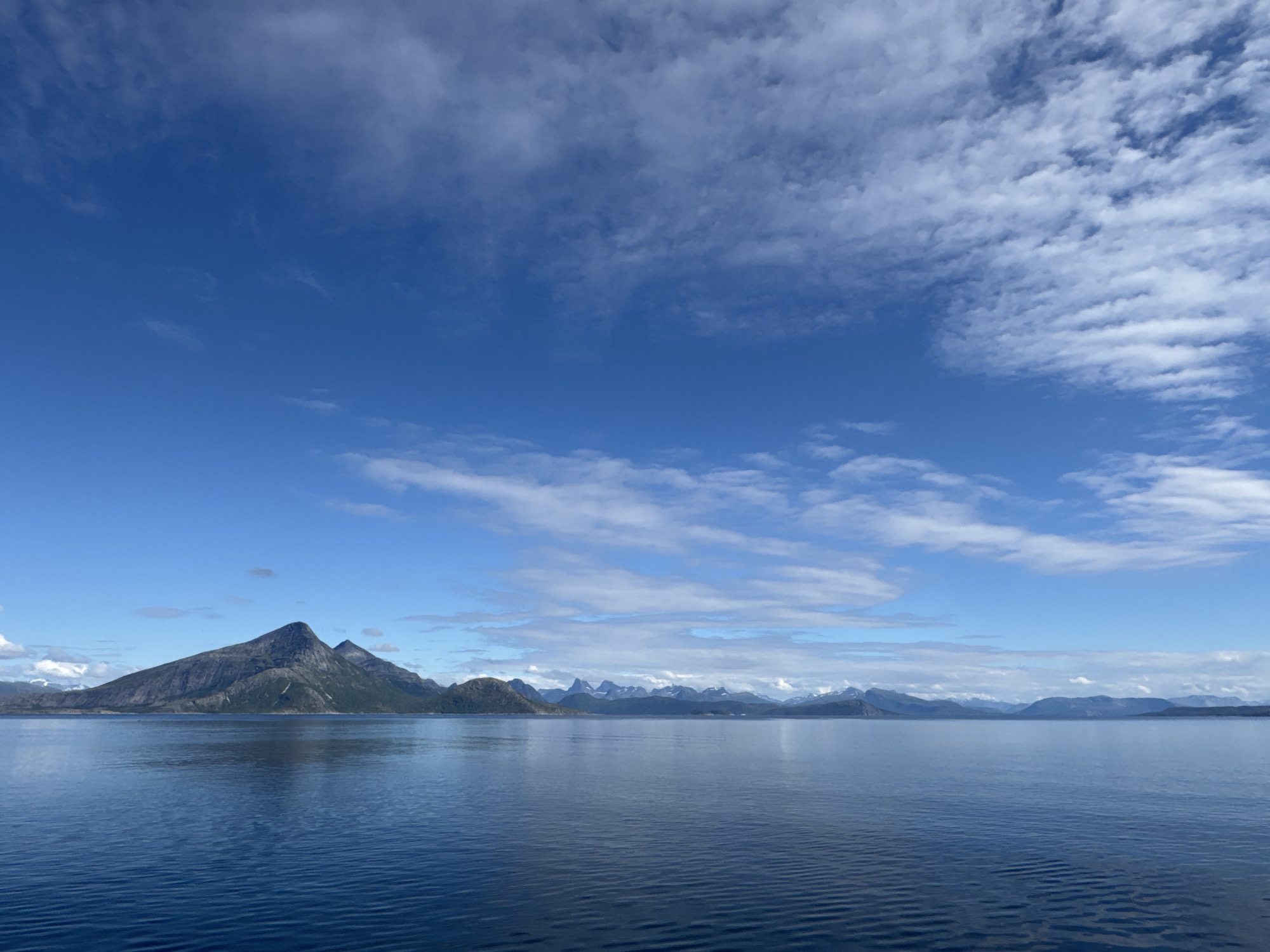 This is a landscape photograph of a body of water with mountains in the background under a bright blue sky with scattered clouds. The foreground consists of calm, deep blue water with small ripples. The middle ground shows a shoreline with dark, rugged mountains. The tallest peak is a prominent, triangular mountain. Further in the distance, a range of smaller, more rounded mountains stretches across the horizon, some with snow-capped peaks. The sky is a vibrant blue, with wispy white clouds, suggesting a clear and possibly sunny day. The lighting indicates it is daytime, likely midday given the brightness. The scene evokes a sense of natural grandeur and tranquility. There is no visible text in the image. The location is likely a fjord or coastal area in Norway, given the mountainous terrain and proximity to water.