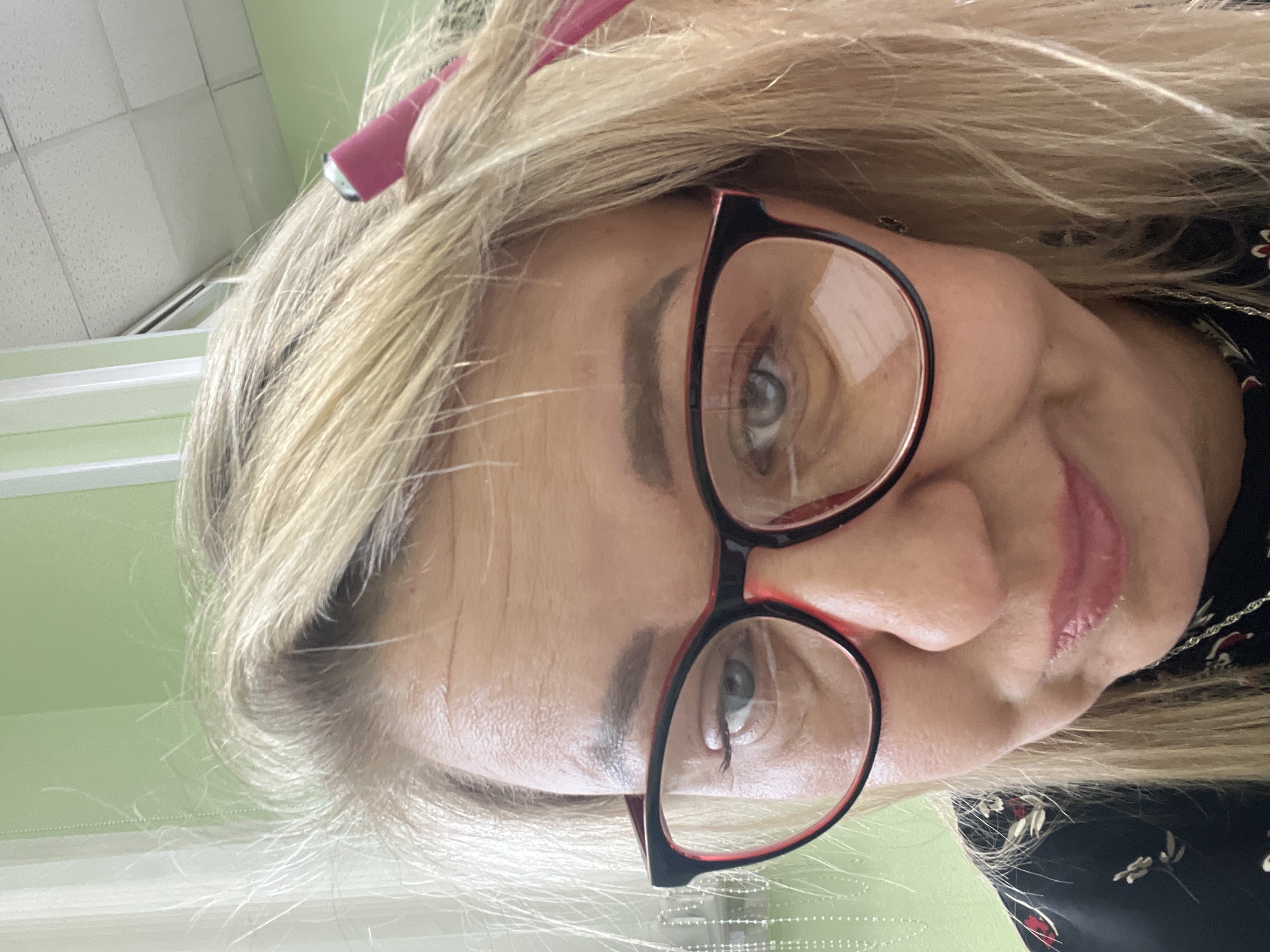 A close-up selfie of a woman with blonde hair, wearing glasses with black and red frames, and a pink hair clip is captured from a slightly tilted perspective. She has fair skin, gray eyes, and is wearing lip gloss. The background shows a light green wall, suggesting an indoor setting. Based on the available information, it is not possible to determine the location within the city of Chaikovsky, Russia.