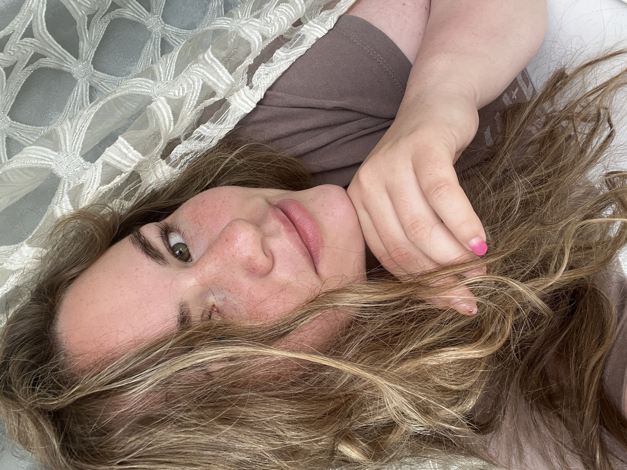 A person with fair skin and freckles is lying down, their head angled towards the camera. They have light brown hair with blonde highlights, some of which is partially covering their face. The person's right hand is near their face, and one fingernail is painted pink. They are wearing a brown t-shirt. A white, patterned fabric is partially covering the person on the left side of the image. The background appears to be a light-colored surface. There is no information visible to identify this image as being in Chaikovsky, Russia.