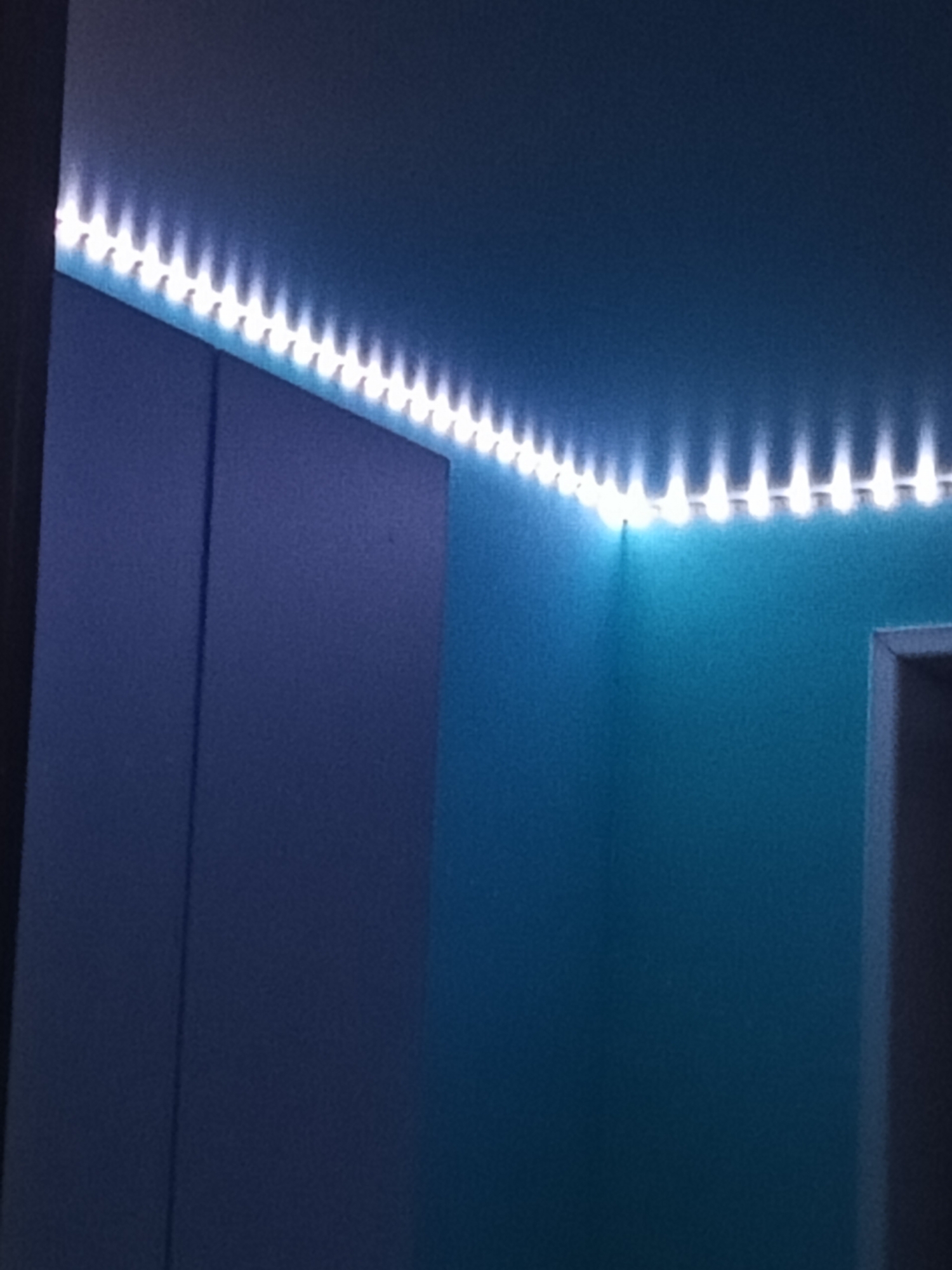 The image displays an interior room, primarily illuminated by a continuous strip of white LED lights affixed to the ceiling, running along the junction of two walls. The ceiling appears light blue-gray. The wall on the right is painted a muted teal or light blue. On the left side of the frame, a large, dark blue or grayish-purple rectangular structure, consistent with a wardrobe or cabinet, extends vertically and features a distinct central seam. A partial view of a doorway with a light-colored frame is visible on the far right. No individuals, specific actions, or interactions are depicted. The visual content provides no forensic indicators to pinpoint a specific location within the city of Chaikovsky, Russia, beyond identifying it as an indoor environment.