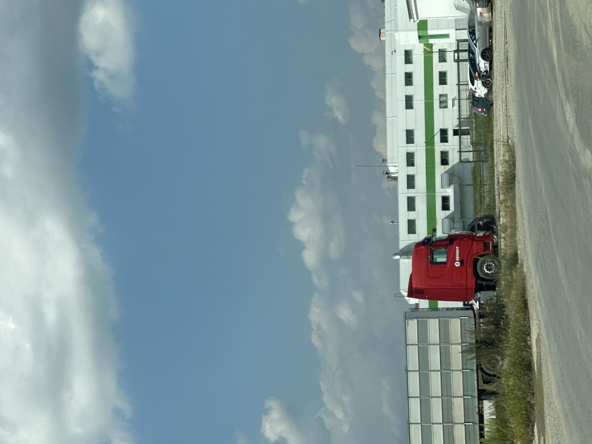 A bright red semi-truck with a white trailer is parked on a dirt lot in front of a white industrial building with green accents. The trailer is a box trailer with multiple horizontal panels. The building has numerous windows and appears to be a factory or warehouse. A line of cars is parked in front of the building. The sky is blue with some scattered white clouds. The overall scene suggests a commercial or industrial area on a clear day.