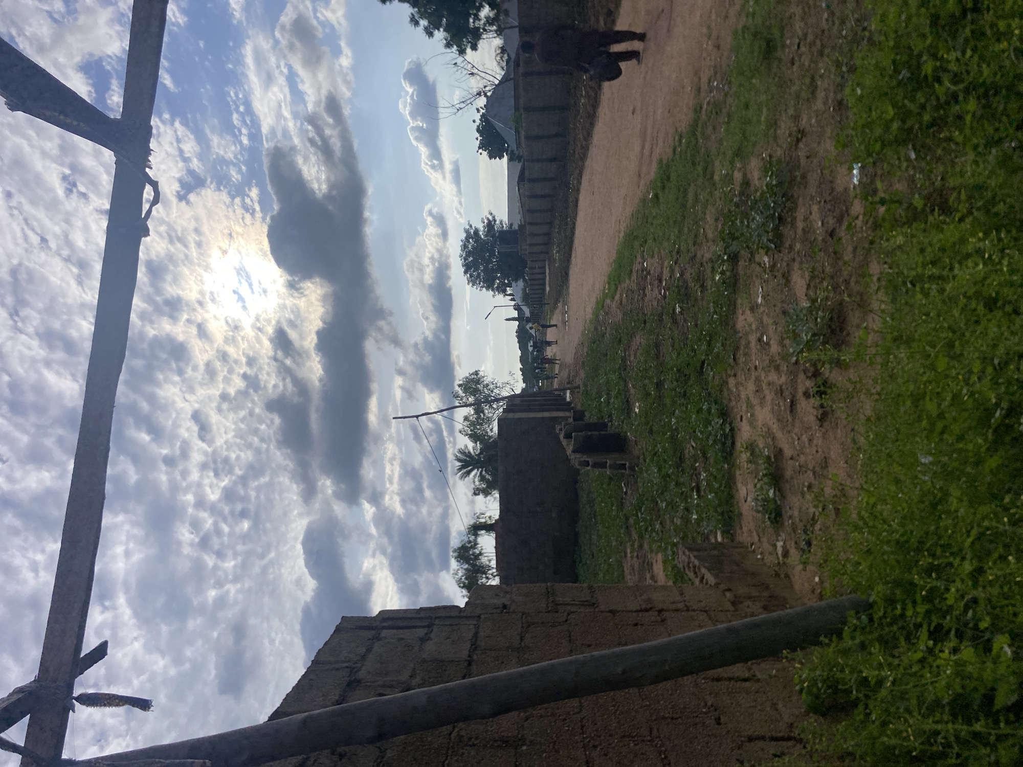 This image depicts an outdoor, semi-urban landscape in Jalingo, Nigeria, captured from a low-angle perspective looking down a dirt path. The upper two-thirds of the frame are dominated by a sky filled with a mixture of altocumulus and stratocumulus clouds, presenting a bright, diffused light source originating from the upper right, where the sun is partially visible behind the cloud cover.
In the immediate foreground, a horizontal wooden beam extends across the top of the frame. From this beam, on the left side, a dark, elongated object resembling a dried corn cob or seed pod hangs vertically. A thick, greyish-brown wooden pole leans diagonally from the lower left towards the center, running parallel to an unfinished block wall constructed of reddish-brown concrete blocks, which occupies the far left of the foreground. The ground in the foreground and along the left edge of the path consists of uneven patches of green vegetation (weeds and grasses) interspersed with exposed light brown earth.
The central feature of the midground is a wide, unpaved dirt path, light brown in color, which extends from the lower center towards the distant horizon. Along the left side of this path, a series of low-rise or incomplete block walls, similar in construction to the one in the foreground, line the route. Further down the path, a small group of approximately five to seven individuals are clustered on the left side, appearing as distant figures. On the right side of the path, an adult male figure, dressed in dark clothing, walks away from the camera, carrying a dark bag in his left hand.
The background features additional sparse tree cover, some with minimal foliage, and what appear to be distant residential structures with light-colored roofs. A utility pole or street lamp is visible on the left side of the path in the mid-distance. The environment suggests an area undergoing development or characterized by rudimentary infrastructure.