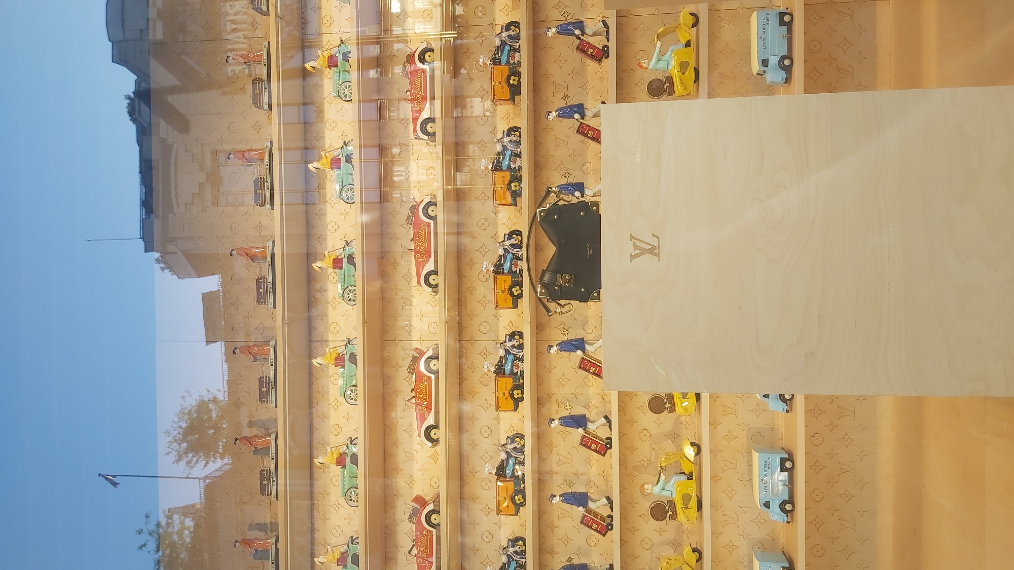 The image shows a display window of a Louis Vuitton store. The display features several rows of miniature vintage vehicles and figurines. In the top row, there are figurines dressed in orange robes driving small wheeled carts carrying what appear to be luggage. Below this, there are several green vintage cars with yellow-clad figurines driving them. The next row down features red and white vintage cars with yellow lettering on their sides. Further down are blue and yellow motorcycles with figures in blue uniforms, some carrying "Louis Vuitton" marked boxes. The bottom rows display blue vans labeled "Louis Vuitton" and yellow scooters with figures. A black Louis Vuitton handbag is also prominently displayed on a pedestal. The background of the display features the Louis Vuitton monogram pattern. The store appears to be located in Paris, France, given the context and the luxury brand. The reflection of the sky and a building with a flag suggest an outdoor setting.