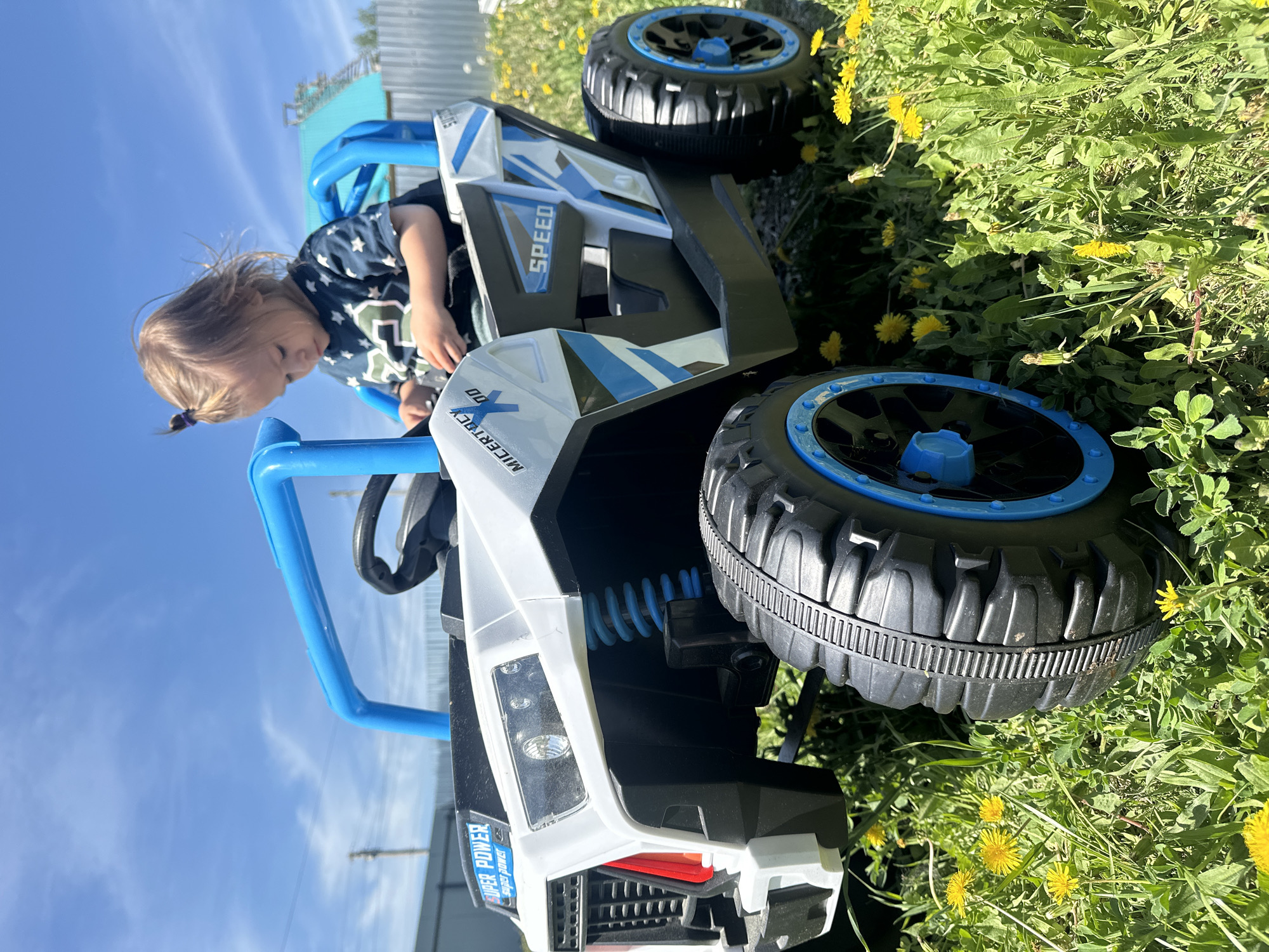 A young child is sitting in a white and blue toy dune buggy on a grassy area covered with dandelions. The dune buggy has black tires with blue rims, and a blue roll cage. The child is wearing a dark shirt with white stars. The sky is blue with some clouds visible in the background. There is a partially visible building or structure made of metal and blue siding. The location is outdoors and is not possible to determine from the image if it is in the city of Kharnavy, Russia.