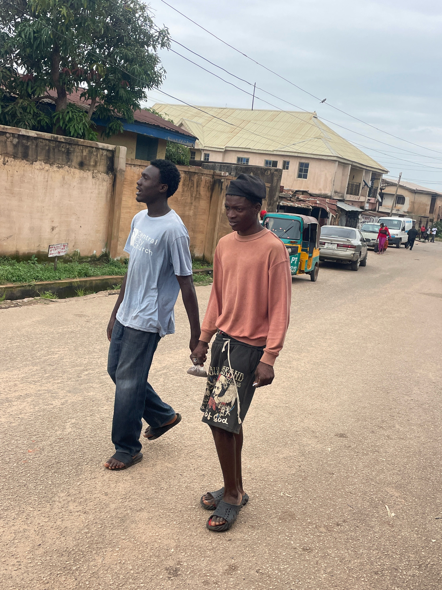 Two young Black males are walking side-by-side on a paved road in a city environment, likely Jos, Nigeria. The male on the left is wearing a light blue t-shirt with "Central City Church" printed on it and dark blue jeans. He is holding the hand of the male on the right. The male on the right is wearing a coral-colored long-sleeved shirt and black shorts with a graphic design. Both are wearing dark-colored open-toed sandals. They are walking away from the camera, towards a street lined with buildings and some vegetation. In the background, there are several vehicles including a green and yellow auto-rickshaw, a silver sedan, and a white van, as well as other pedestrians. The sky is overcast. The road surface is asphalt with visible aggregate. A beige concrete wall runs along the left side of the road.