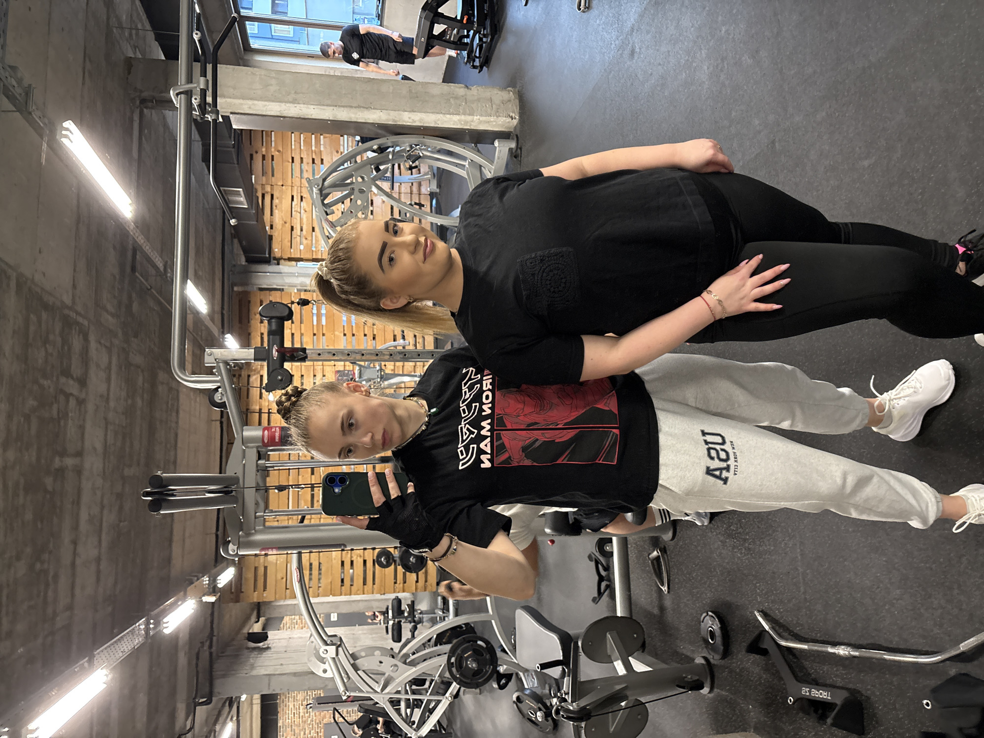 The image shows two young women posing for a selfie in a gym setting. The gym has a variety of exercise equipment visible, including weight machines, bars, and plates. There is also exposed brick and palleting on some of the walls.
The woman in the foreground is holding a smartphone and taking the picture. She is wearing a black t-shirt, grey sweatpants, white shoes, and workout gloves. Her hair is styled in a bun. The woman in the background is wearing a black t-shirt, black leggings, and black sneakers. Her hair is in a ponytail.
The location can not be determined to be Sofia, Bulgaria from the content of this image.