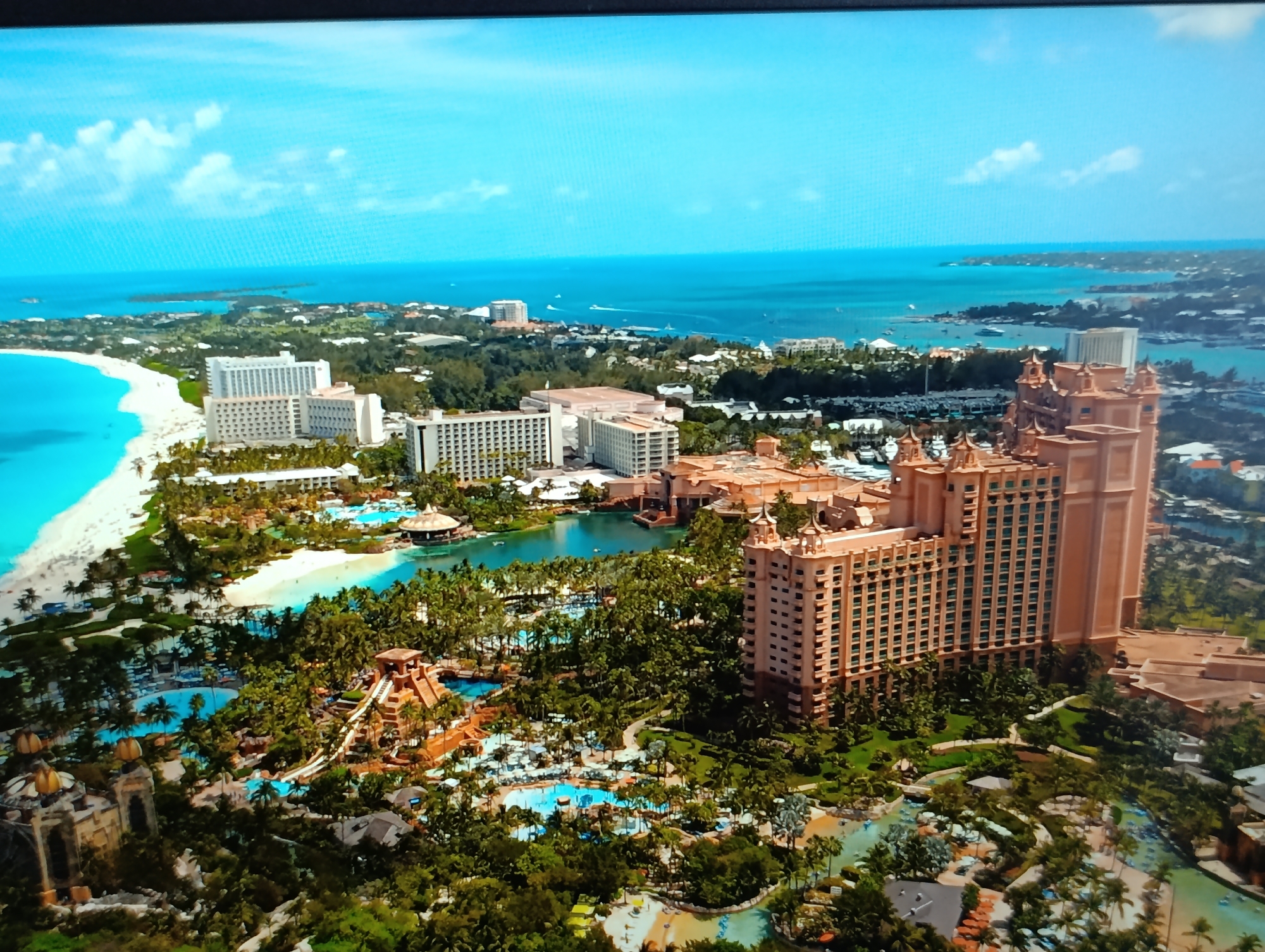 This is an aerial photograph of a coastal resort. The resort features a large, peach-colored hotel with multiple floors and a distinctive architectural style, likely designed to evoke a sense of historical grandeur. Surrounding the hotel is an expansive complex of swimming pools, water slides, and lush tropical landscaping with numerous palm trees. A white sandy beach with turquoise water is visible along the coastline. In the background, the ocean stretches to the horizon, dotted with several boats. The sky is a bright blue with scattered white clouds. The location is not in Dubăsari town council, Moldova; it appears to be a tropical resort destination.