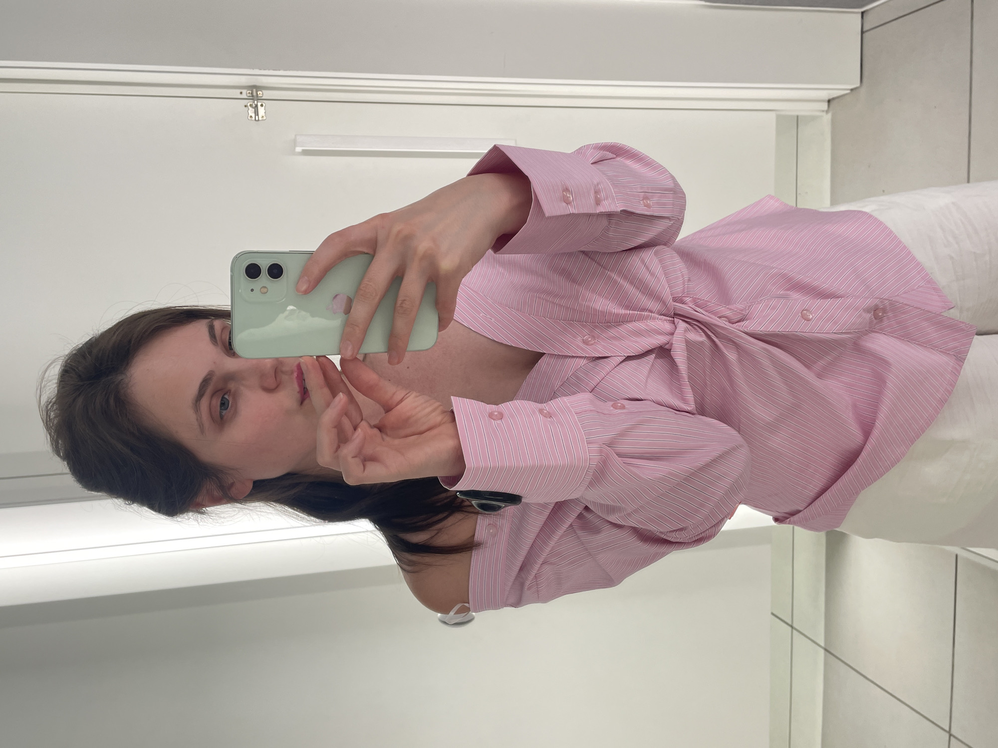 An overhead shot shows a woman taking a mirror selfie in a fitting room. She is wearing a pink and white striped off-the-shoulder shirt with a knot in the front and white pants. She is holding a light green iPhone in her right hand, and her left hand is near her face. The fitting room has white walls, a white door with a silver latch, and white tiled flooring. The lighting appears to be overhead fluorescent lighting.