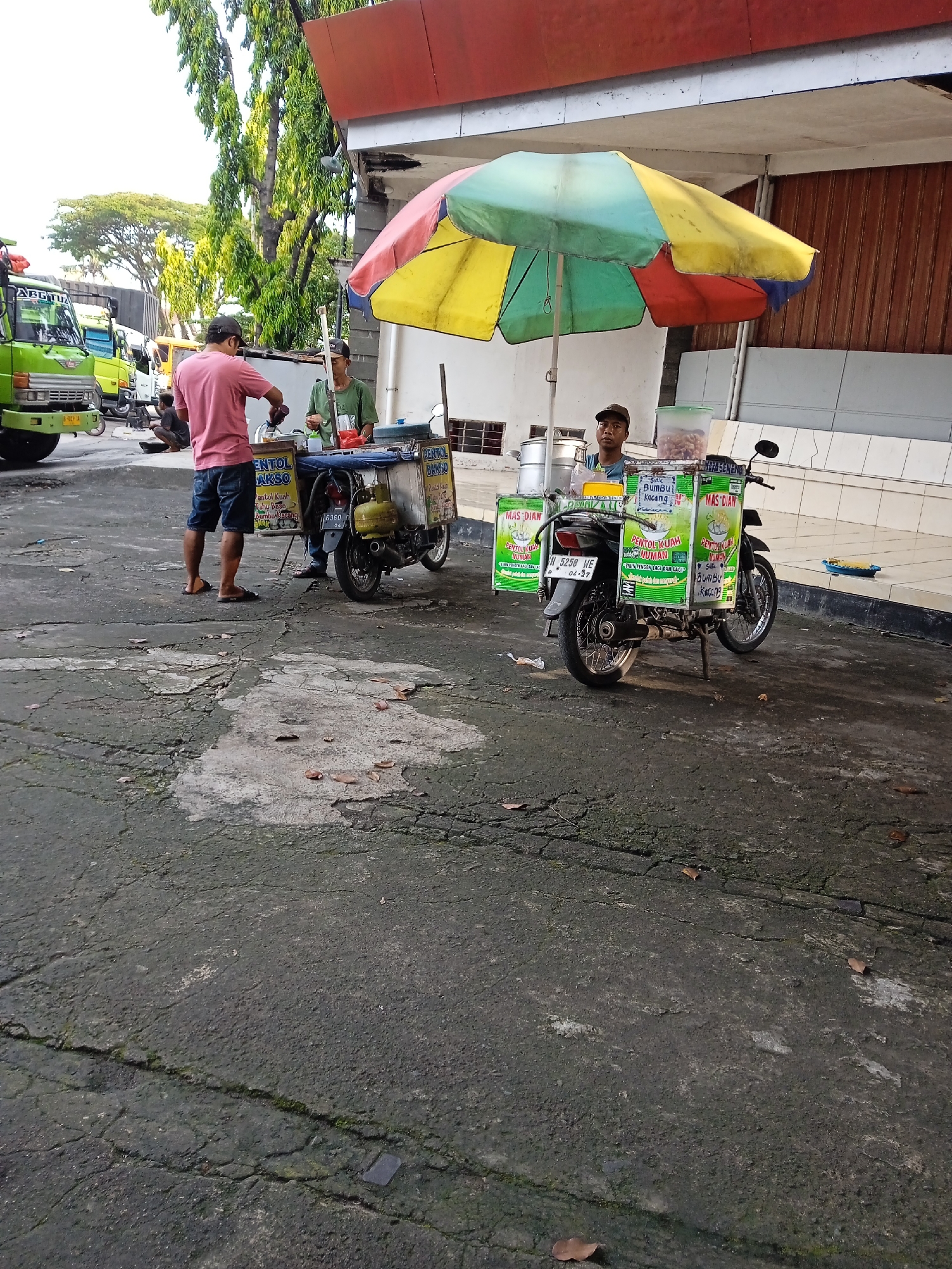 The image captures a street scene in Wonoketingal, Indonesia. Two mobile food vendors, each operating from motorcycles equipped with side-mounted stalls and colorful umbrellas, are present. One vendor, wearing a green shirt and cap, attends to a customer in a pink shirt and blue shorts. The stalls advertise "PENTOL AKSO" and "MAS DIAN," likely local food offerings. The area appears to be a paved lot with cracks and leaves scattered on the ground. In the background, several trucks are parked, adding to the bustling atmosphere. The weather is bright and sunny, casting shadows across the pavement.