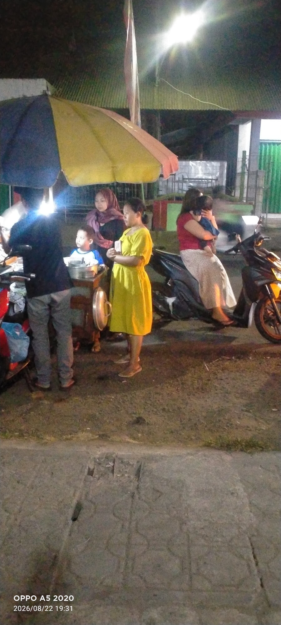 This image captures a scene at night in Kedung, Indonesia, near what appears to be a street food vendor. A yellow and blue umbrella shades a group of people. In the foreground, a person in grey pants stands near a cart with a metal pot. To their left, a young boy in a blue and white shirt looks towards the cart. To the right of the boy, a woman in a yellow dress holds what appears to be a small white object. Behind her, a woman in a purple hijab stands, looking towards the cart. Further to the right, another woman in a red shirt is seated on a motorcycle, holding a baby. The environment is dimly lit, with artificial lights illuminating the scene. The ground is paved with concrete. The timestamp overlay indicates the image was captured on August 22, 2026, at 19:31.