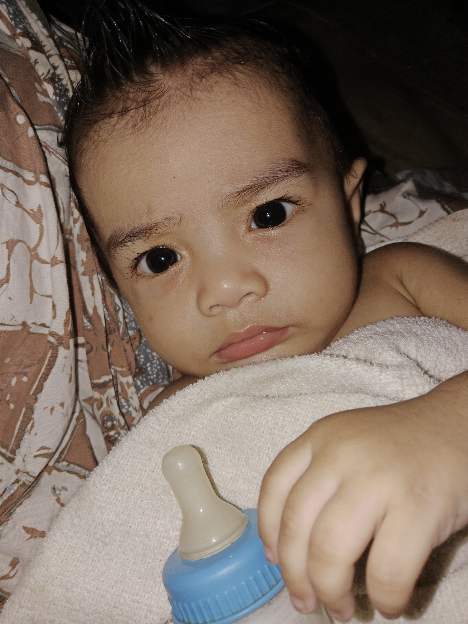 A close-up, slightly angled shot shows a baby lying down with a blue and white baby bottle. The baby, with dark hair, is looking directly at the camera with wide, dark eyes and slightly parted lips. The baby is wrapped in a white towel, and a patterned fabric, possibly a blanket or bedding, is visible behind the baby's head and to the left. The lighting is dim, creating shadows on the right side of the image. The scene appears to be indoors.