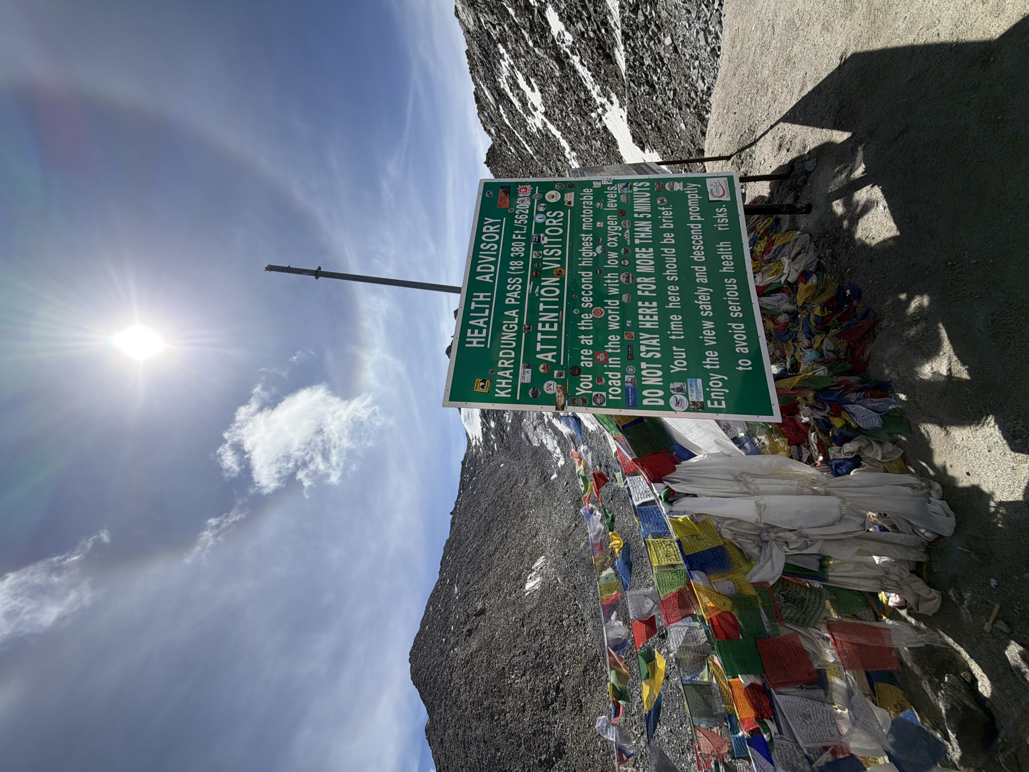 The image shows the Khardungla Pass in Leh, India, which is at an altitude of 18,380 feet (5620 meters). A green sign displays a health advisory for visitors, warning them about the low oxygen levels and advising them to limit their stay to five minutes to avoid health risks. The pass is decorated with colorful prayer flags, which are a common sight in Buddhist regions. In the background, a rocky mountain slope with patches of snow can be seen, and the sun is shining brightly in a blue sky with some clouds, creating a halo effect. The overall scene is a rugged, high-altitude landscape typical of the Himalayas.